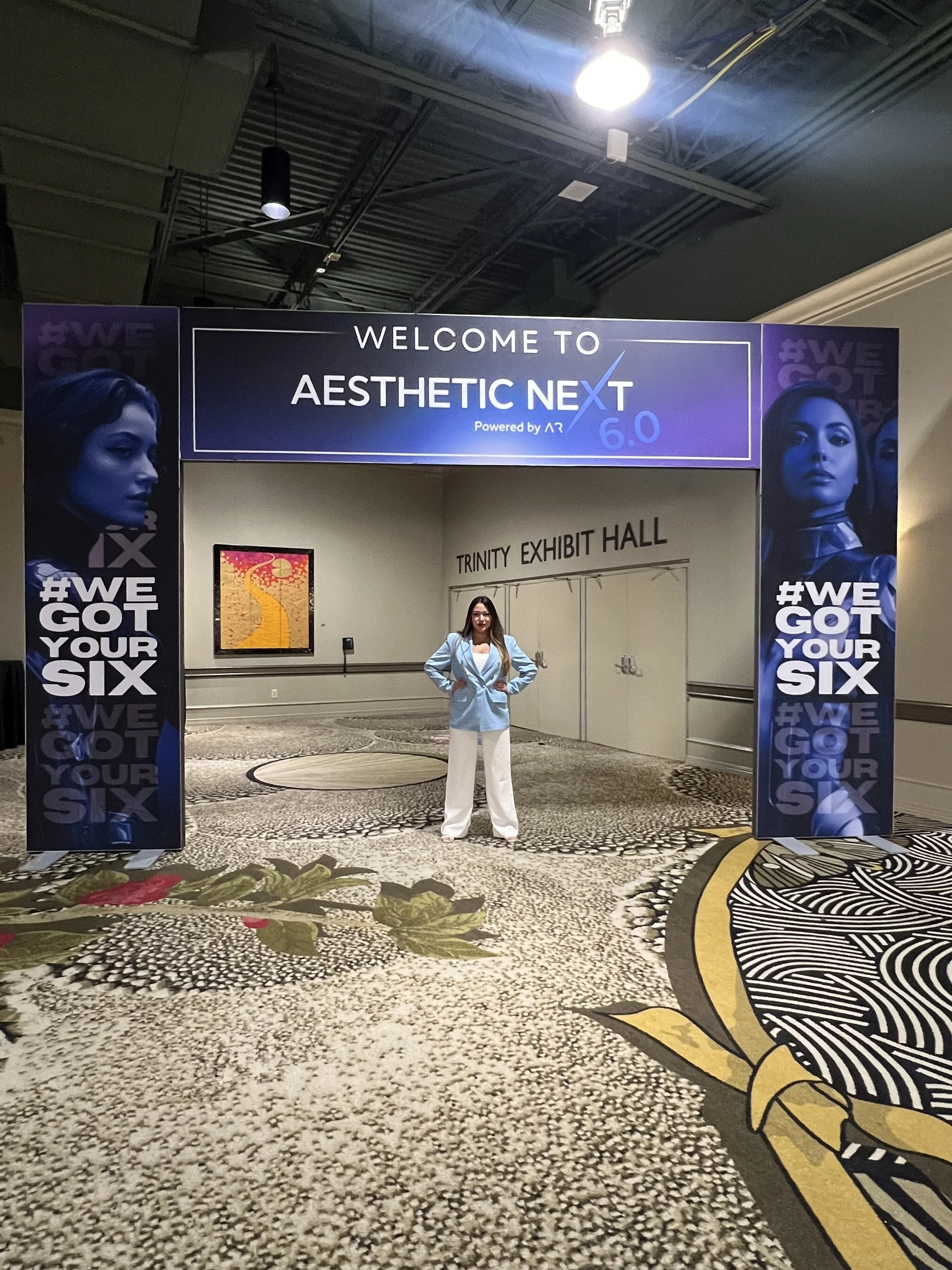 A woman in a light blue blazer and white pants standing under a large purple sign that reads "Welcome to Aesthetic Next 6.0," with French text "Powered by AR." The sign features images of women on the sides and social media hashtag "#WegotYourSix." T