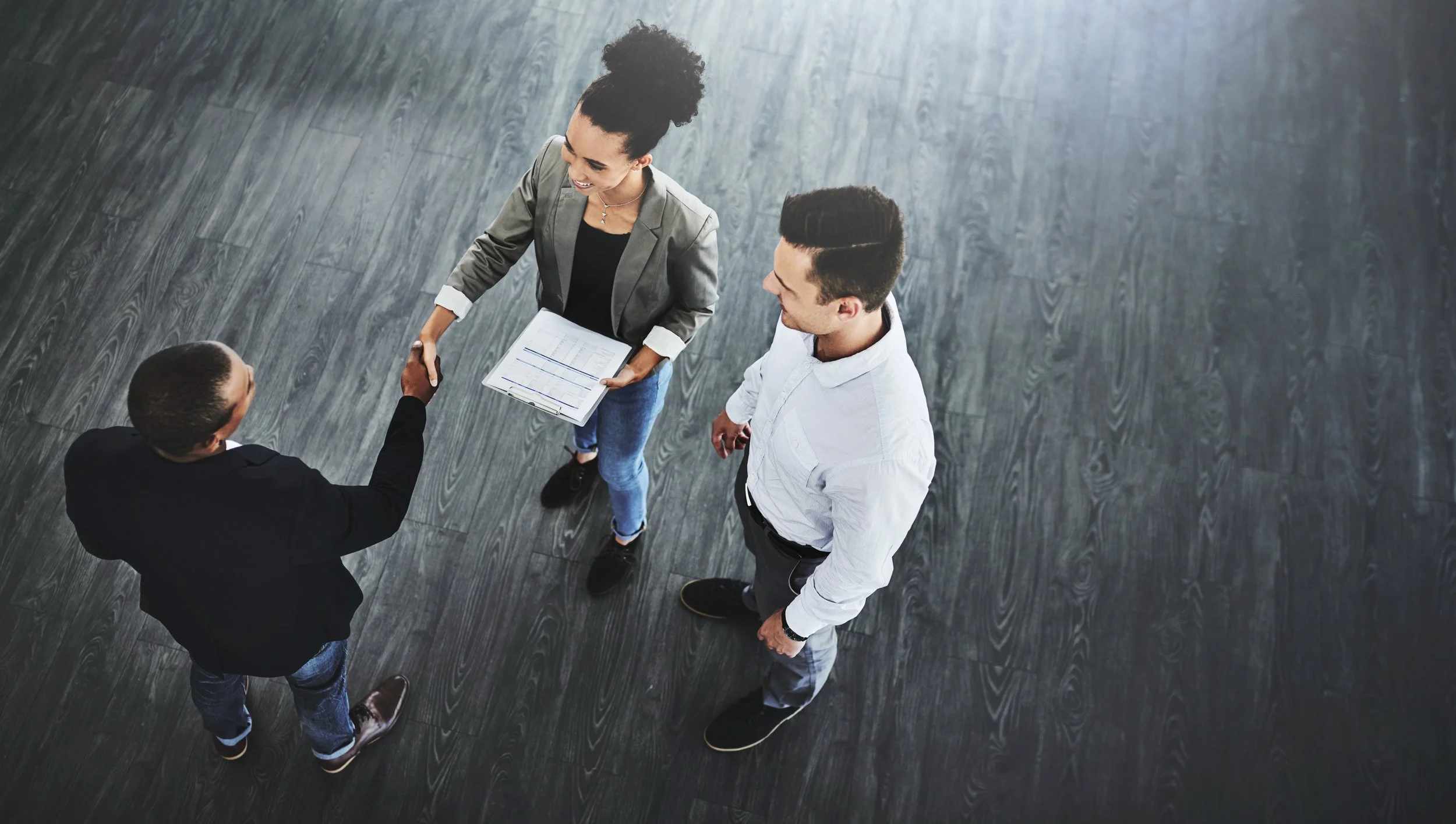 Three people standing on dark wooden floor in a business setting. A woman in a gray blazer and holding documents shakes hands with a man in a black suit, while another man in a white shirt observes.