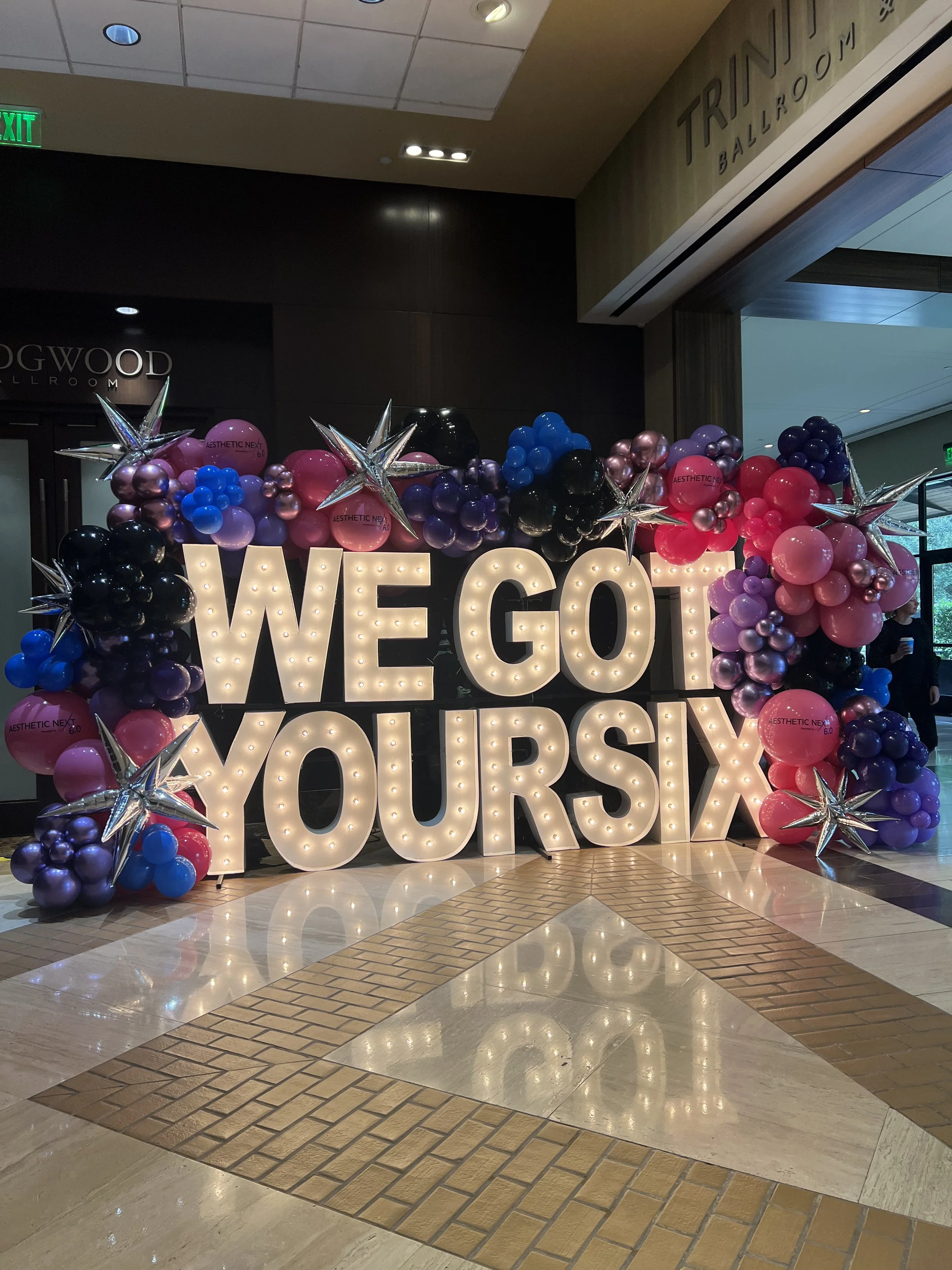 Decorative display with large illuminated sign saying "WE GO YOURS" surrounded by purple, pink, black, and blue balloons and silver star-shaped balloons, located in a mall or event space.