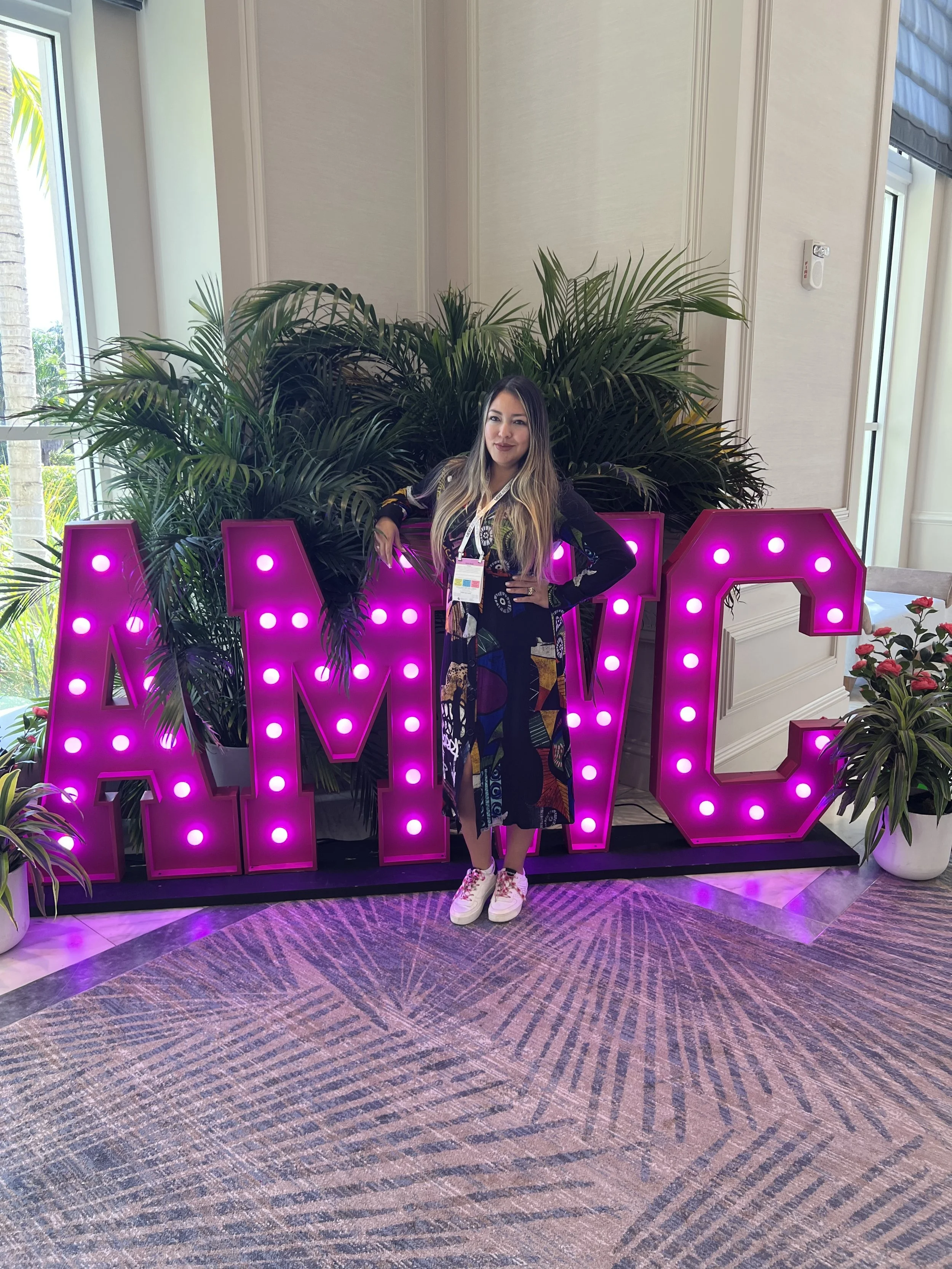 Woman wearing a colorful dress and white sneakers standing in front of large illuminated pink letters spelling 'AMG' with indoor plants behind her.