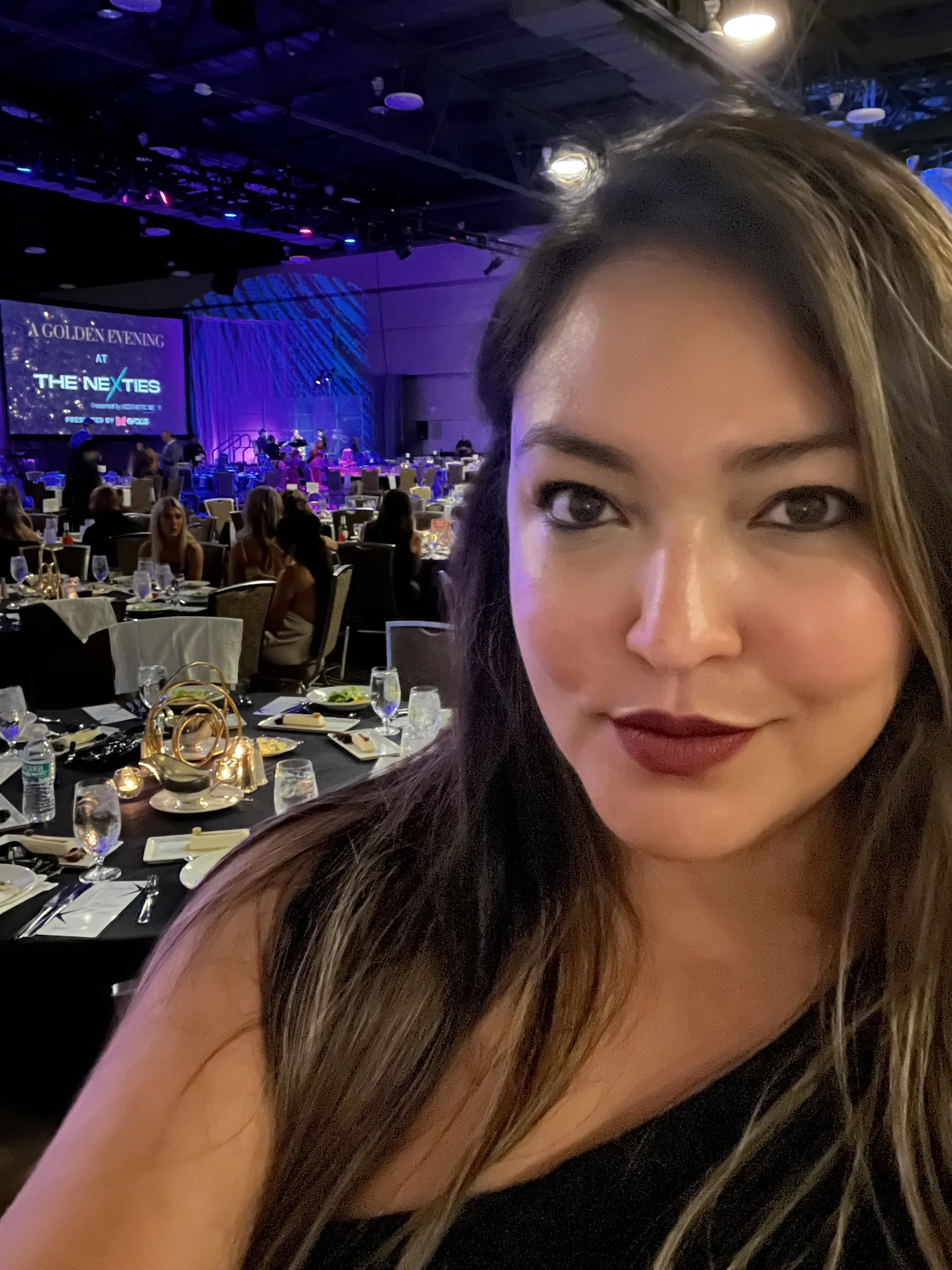A woman with long, dark hair and makeup posing at a formal dinner event. The background features tables with dining setups and a stage with a large screen displaying 'A Golden Evening at The NexTees.'