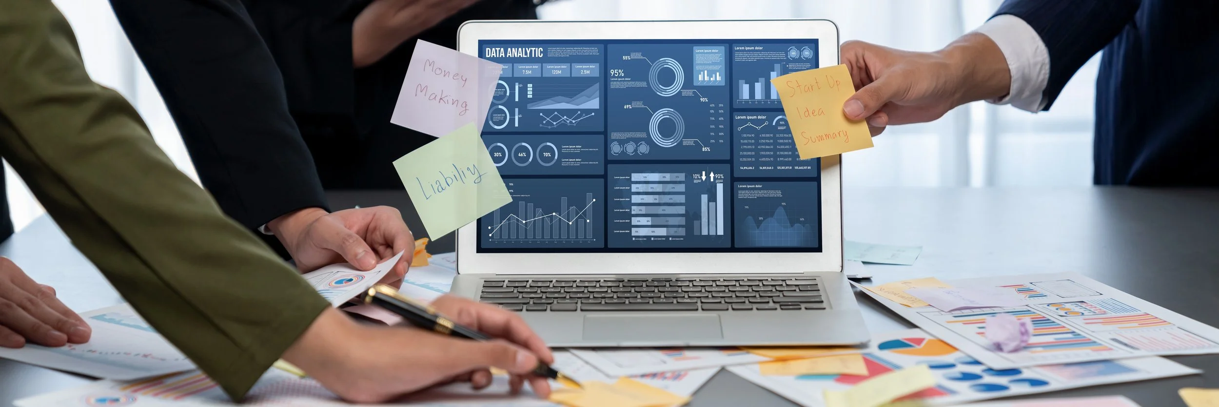 People working on a data analysis project with a laptop showing charts and graphs, surrounded by papers and sticky notes with notes like 'Money Making,' 'Liability,' and 'Start Up Idea Summary'.
