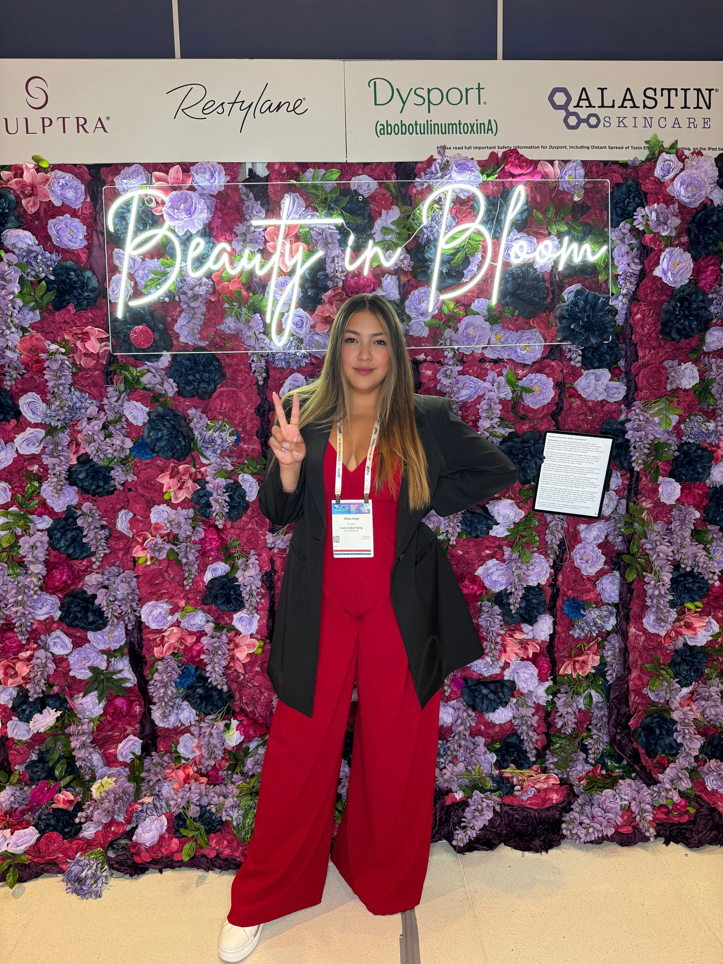 A woman with long hair, dressed in red pants and a black jacket, posing in front of a floral backdrop with a neon sign that says 'Beauty in Bloom', making a peace sign with her right hand.