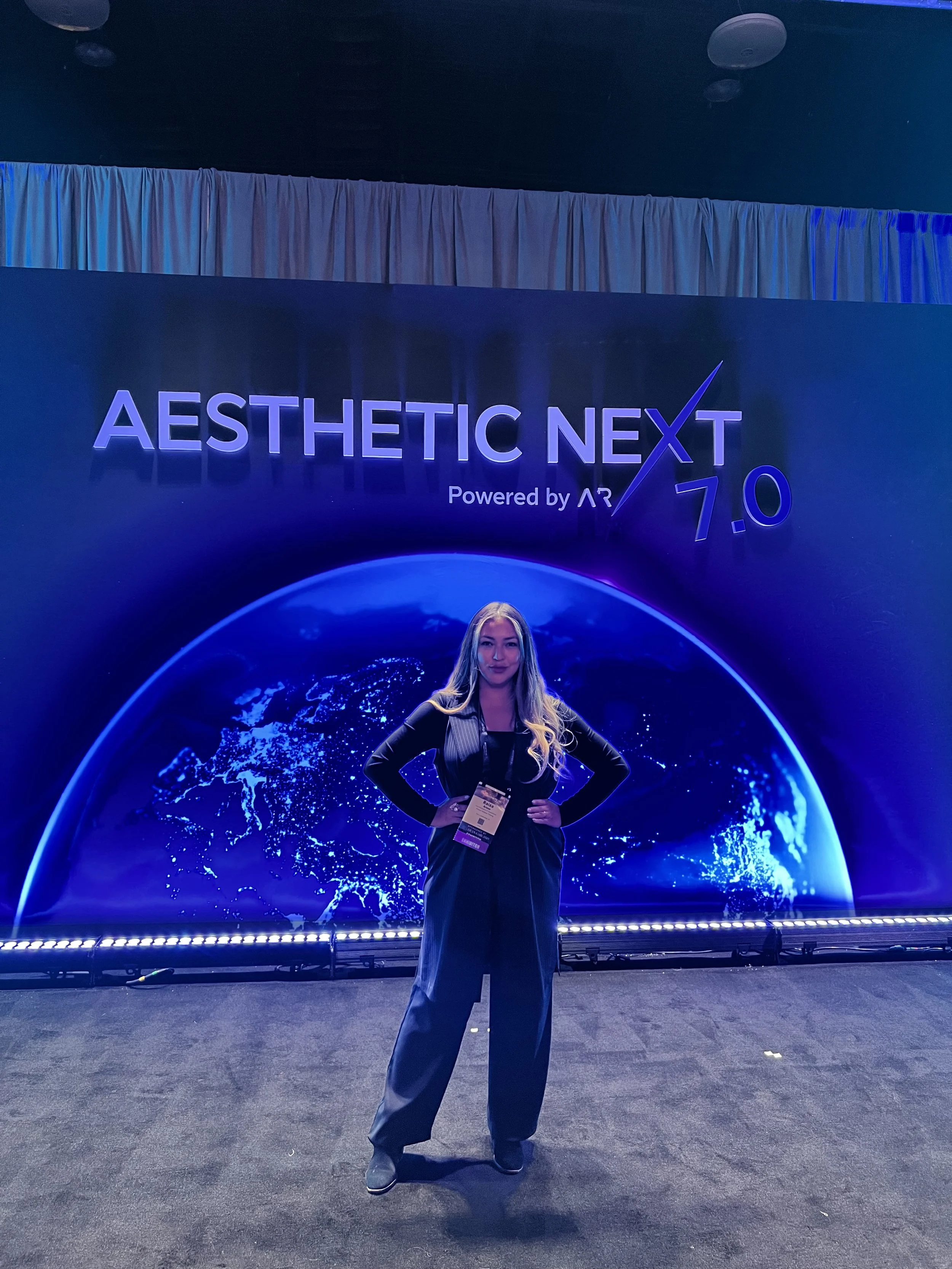 A woman standing in front of a large display screen at a technology event. The display reads 'Aesthetic Next Powered by AR 7.0' with a digital Earth graphic behind her. The woman is wearing a black outfit, has long blonde hair, and is posing with her