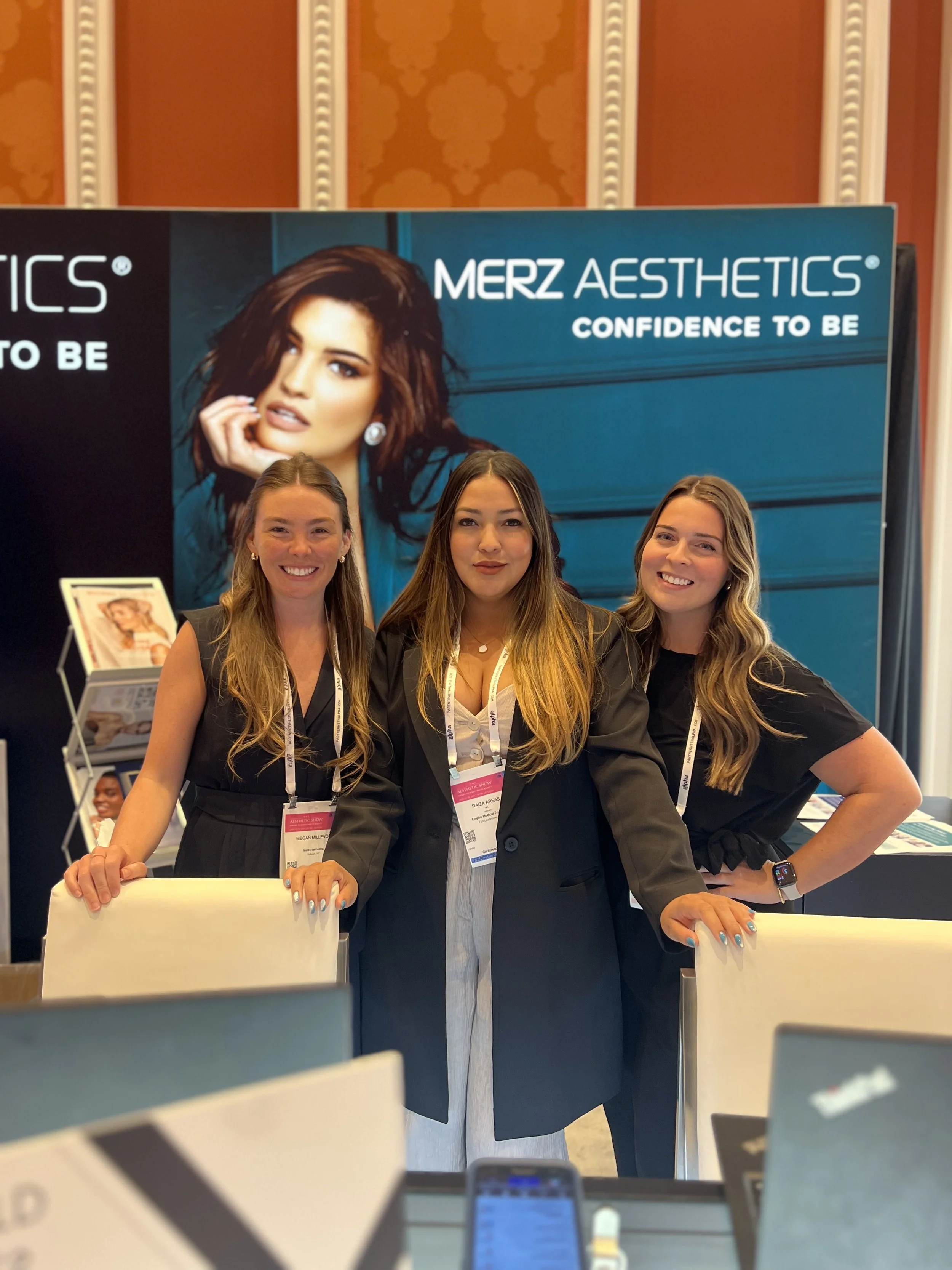 Three women standing at a booth with a blue backdrop that has the text 'MERZ AESTHETICS' and 'CONFIDENCE TO BE'. They are wearing business attire and name tags, smiling for the photo.