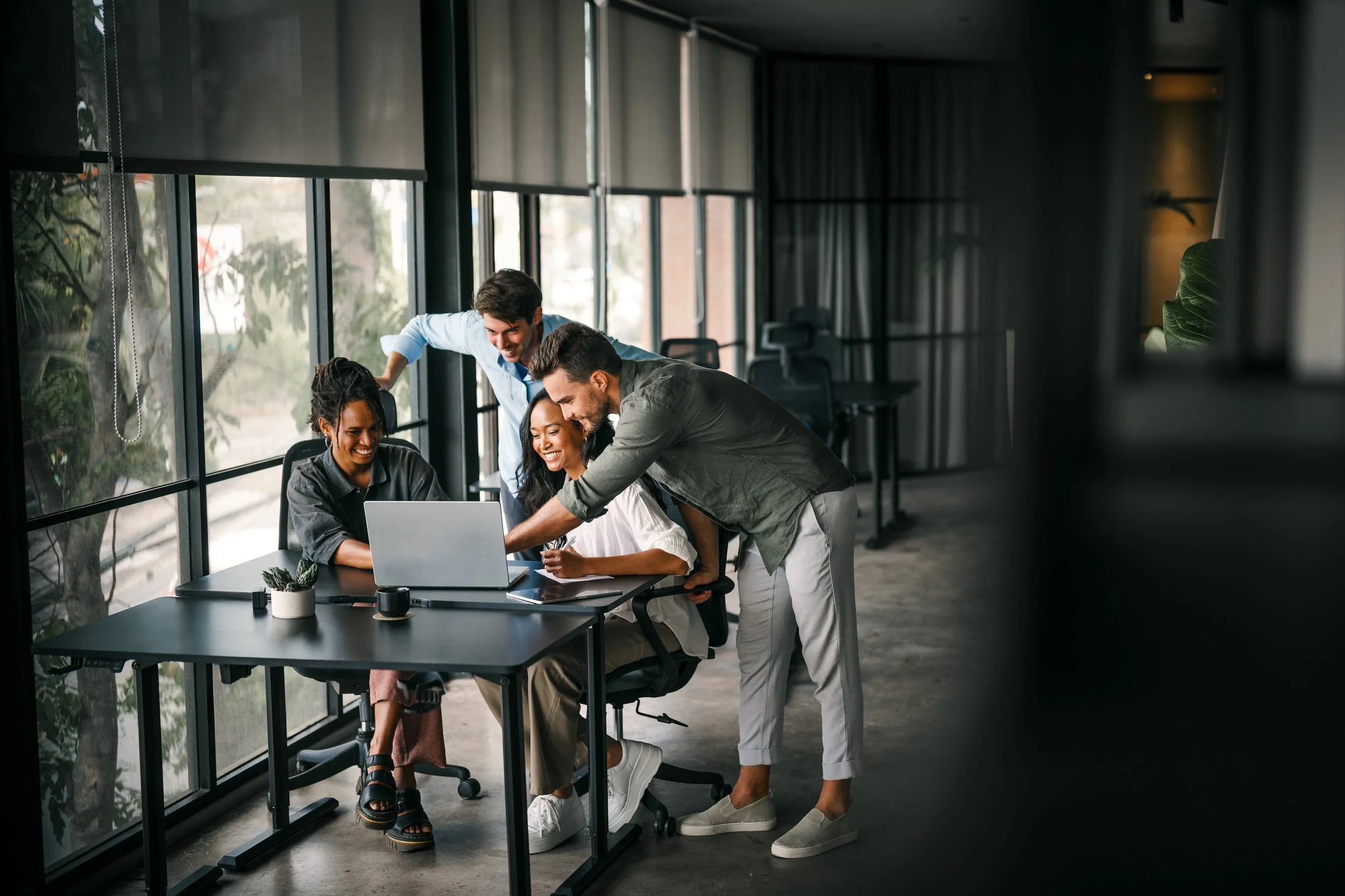 A diverse group of five young professionals, three women and two men, gathered around a laptop in a modern office with large windows. They are smiling and collaborating, with two people standing and leaning over the seated individuals, who are engaged with the laptop. The office has a sleek, minimalist design with a table, small plant, and coffee cups in the foreground.