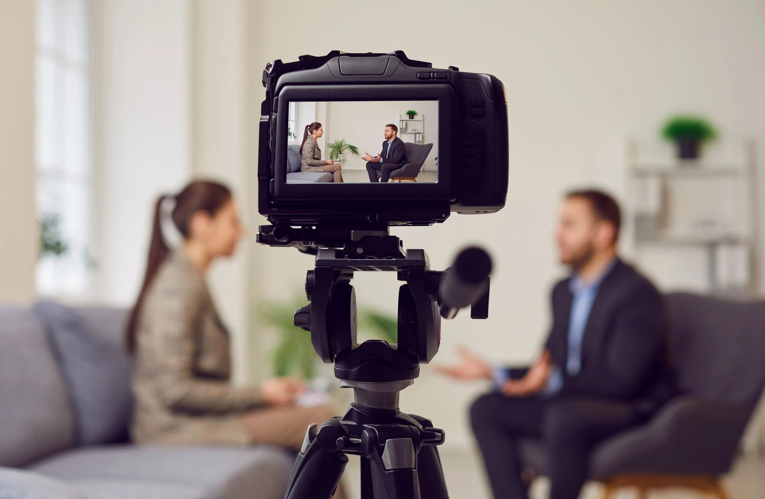 Camera capturing a video interview of a woman and a man seated in a room with white walls and plants in the background.