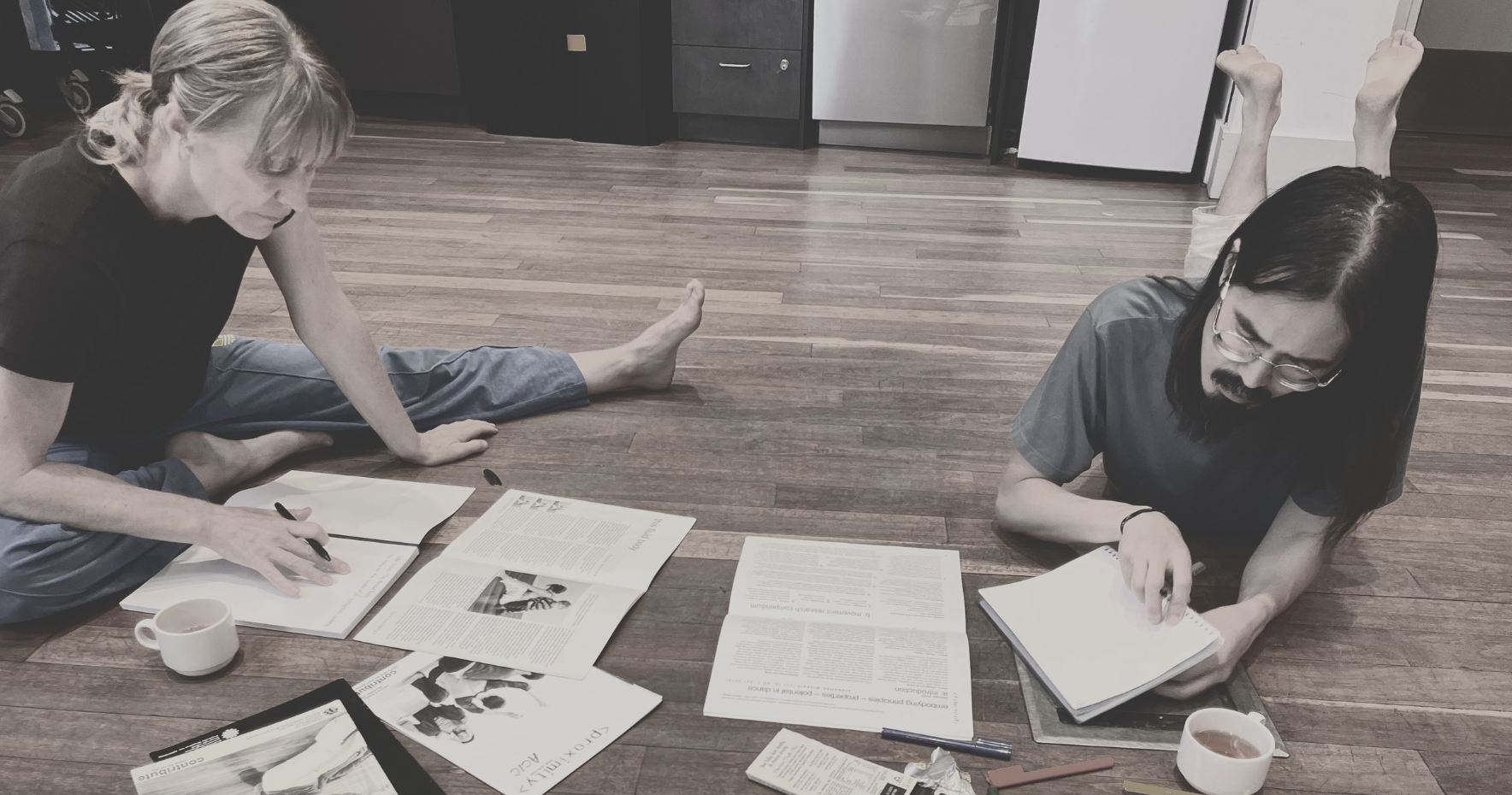 Two people lie on a hardwood floor, reading and working with papers, magazines, and notebooks. One person with long dark hair and glasses is on the right, while the other with short blonde hair is on the left, with their legs stretched out. There are cups, pens, and other reading materials around them.