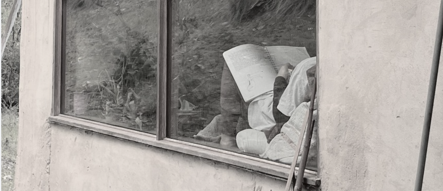 Person sitting on the floor reading a large book inside a room, viewed through a window with a wooden frame.