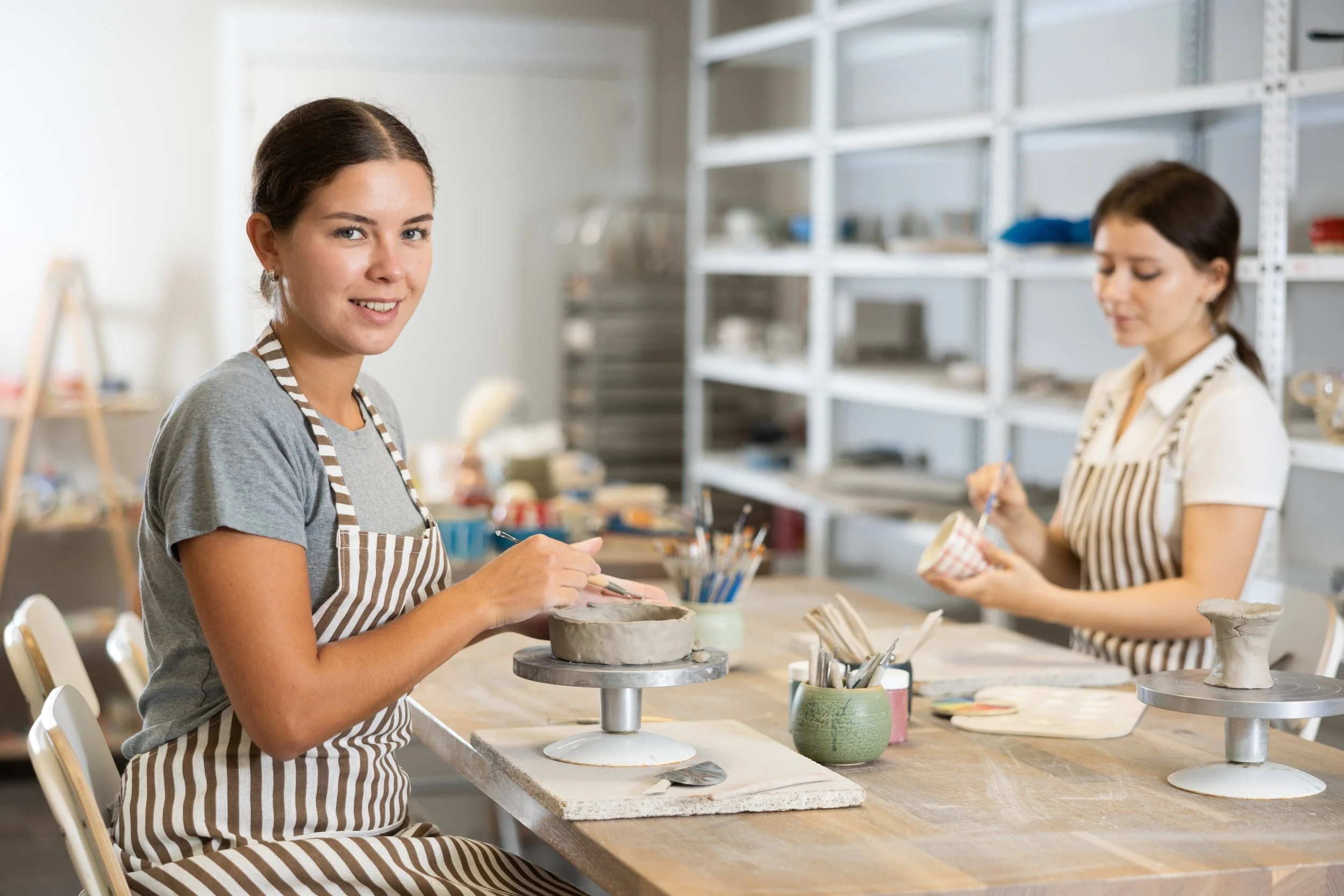 Two women working on pottery at a studio. The woman in the foreground is smiling, wearing a striped apron, and working on a pottery wheel. The woman in the background is focused on shaping clay.