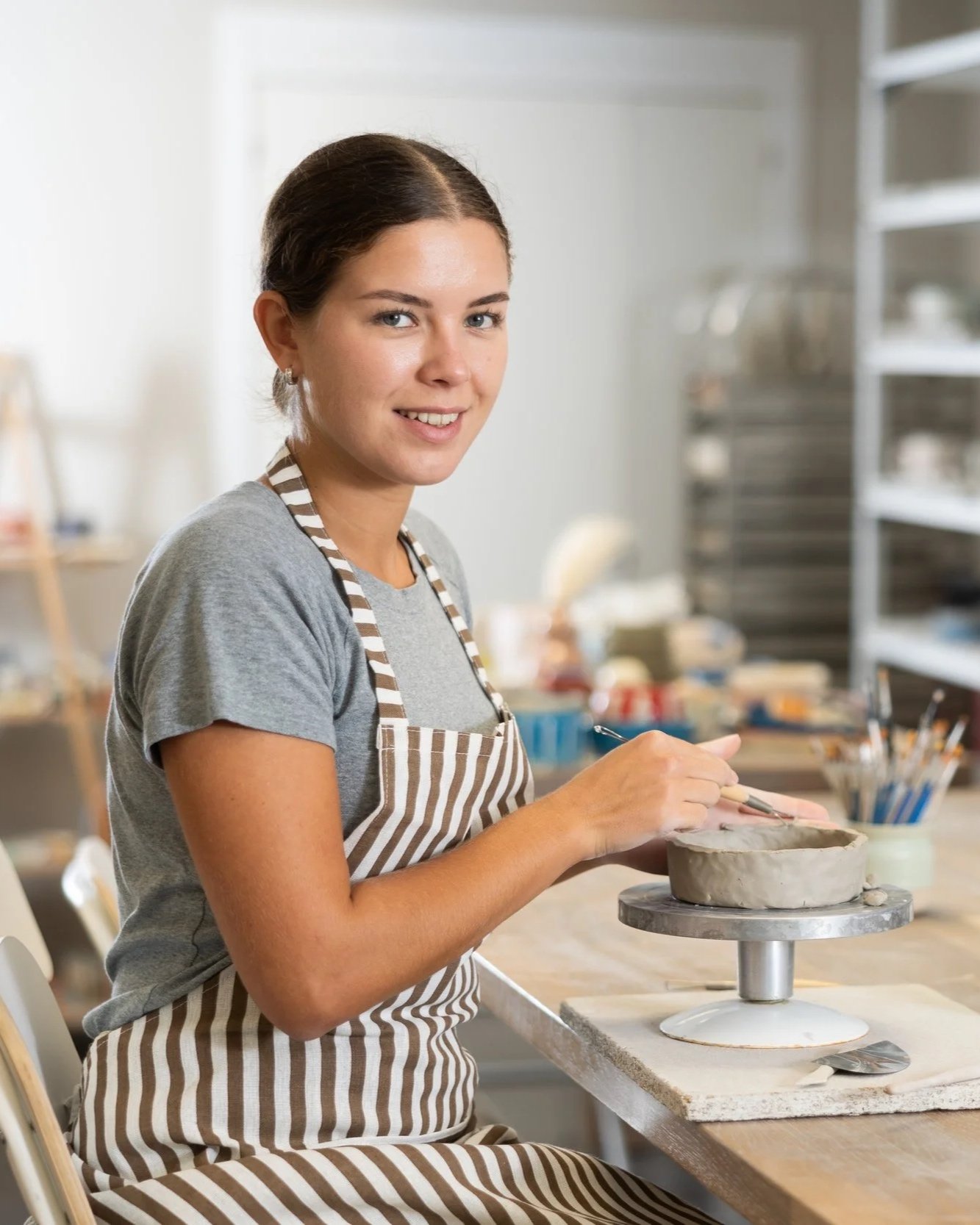 A young woman with dark hair tied back, wearing a gray T-shirt and a striped apron, is working on a pottery wheel in a ceramic studio, shaping a piece of clay.