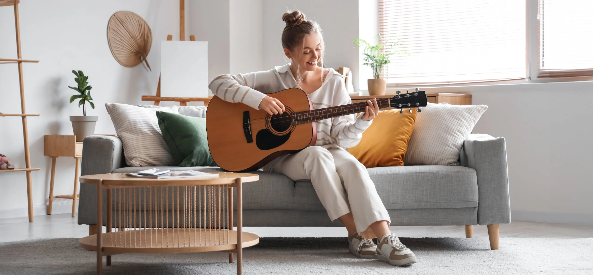 A woman sitting on a gray sofa, playing an acoustic guitar, in a bright living room with modern decor, pillows, and houseplants.