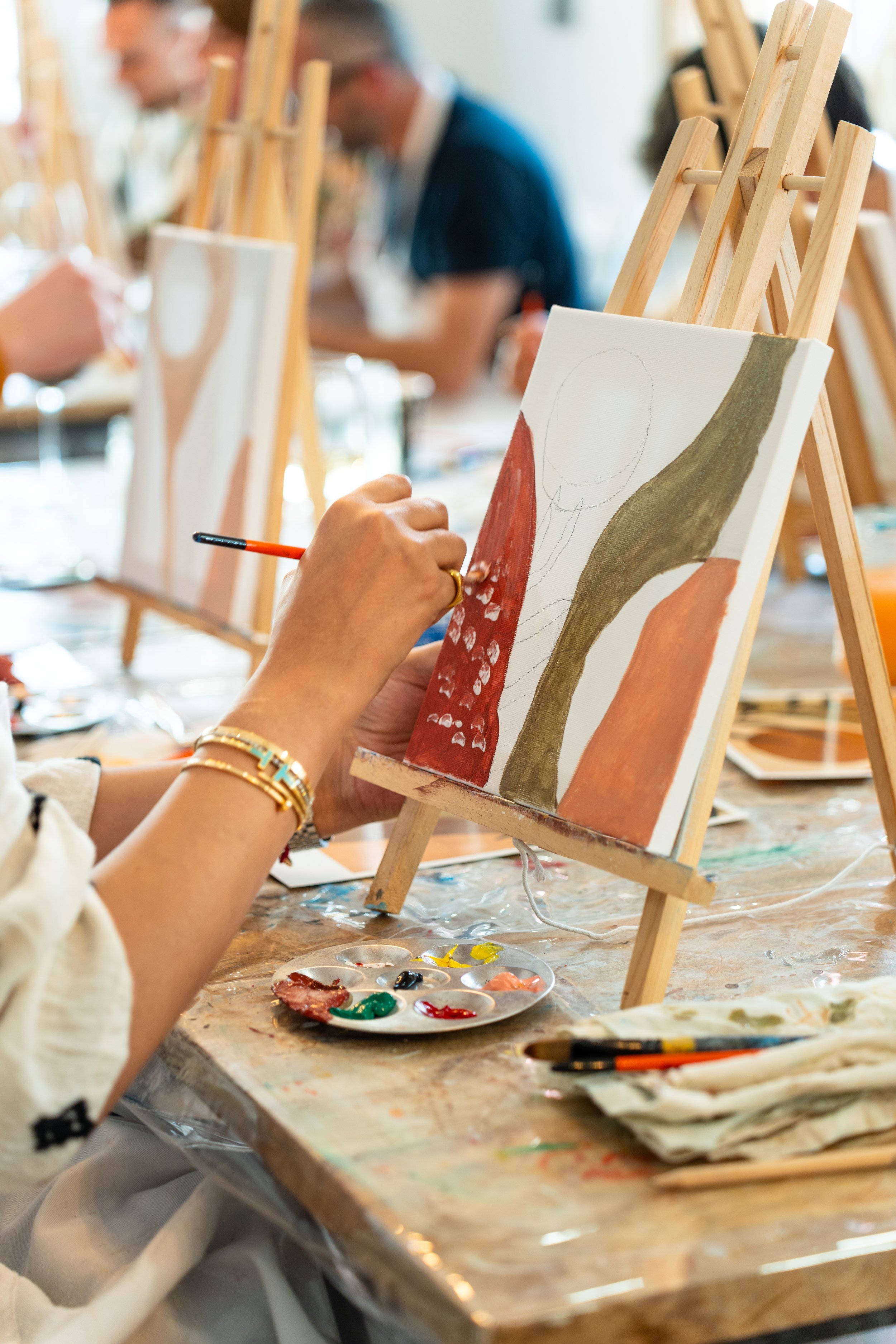 A person painting an abstract artwork on a canvas with a small brush, surrounded by painting supplies and other easels in a bright, lively art studio.