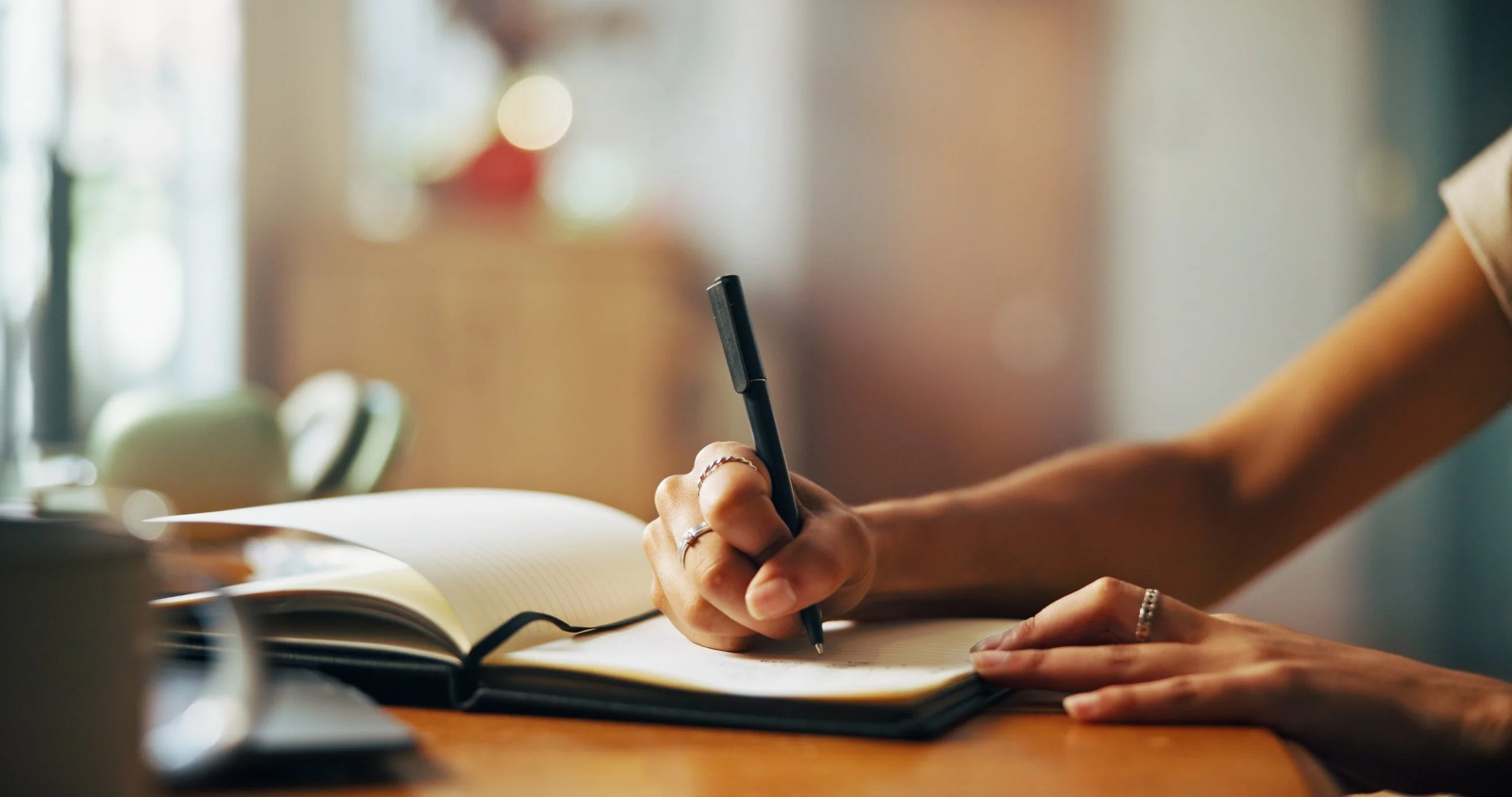 A person writing in a notebook with a black pen, sitting at a desk with a blurred background of a window with natural light.