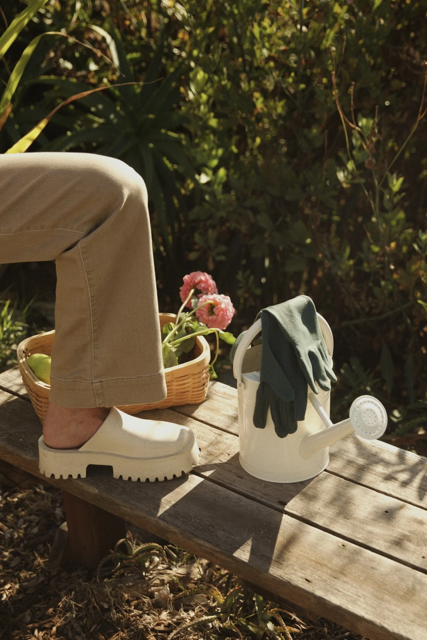 A beige pair of shoes with white soles on a wooden picnic table. Next to the shoes is a small white watering can with a pair of green gloves hanging on its handle. In the background, there are plants with green leaves and pink flowers, illuminated by