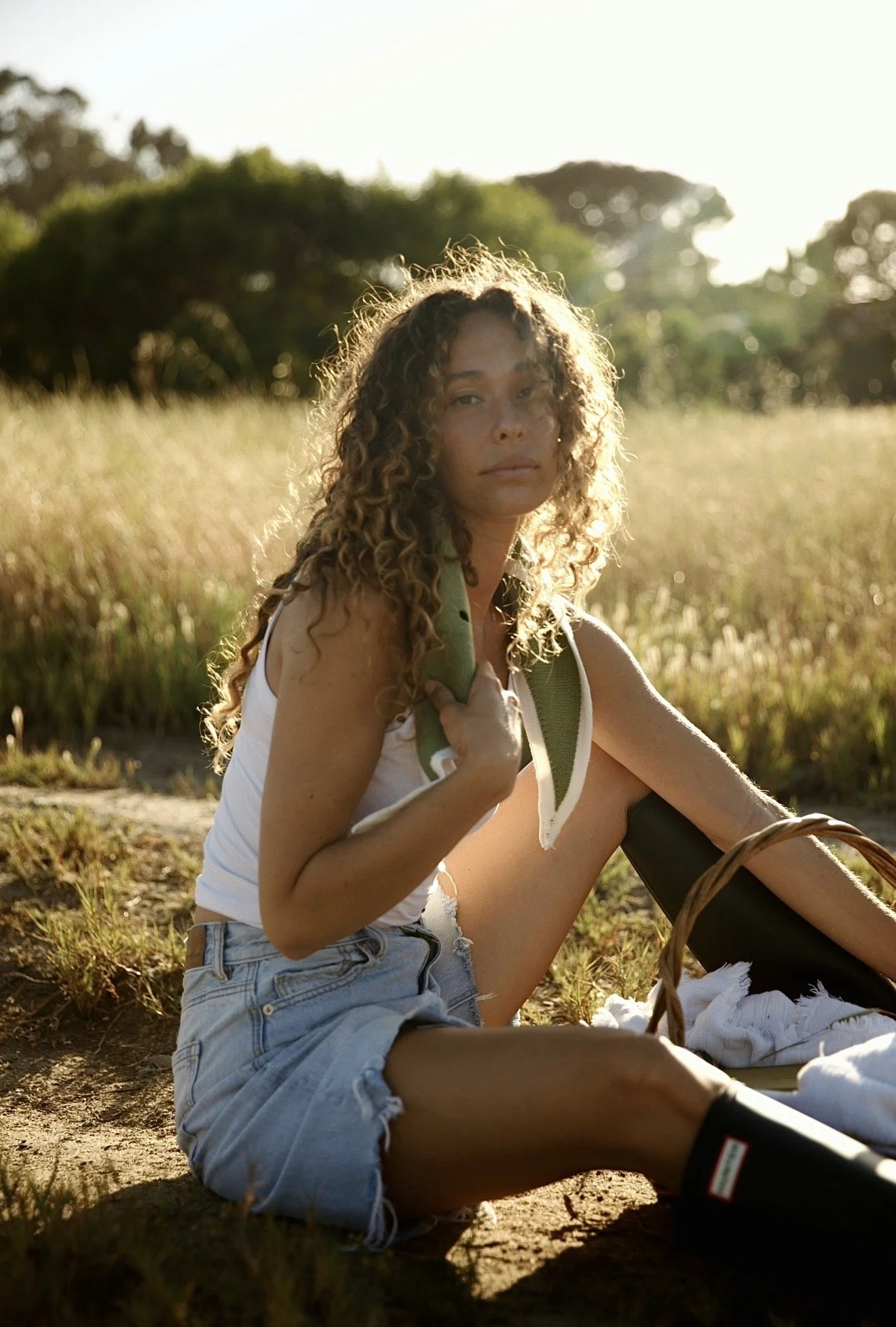 A young woman with curly hair sitting outdoors in a field during sunset, holding a green water bottle, wearing a white tank top and denim shorts.