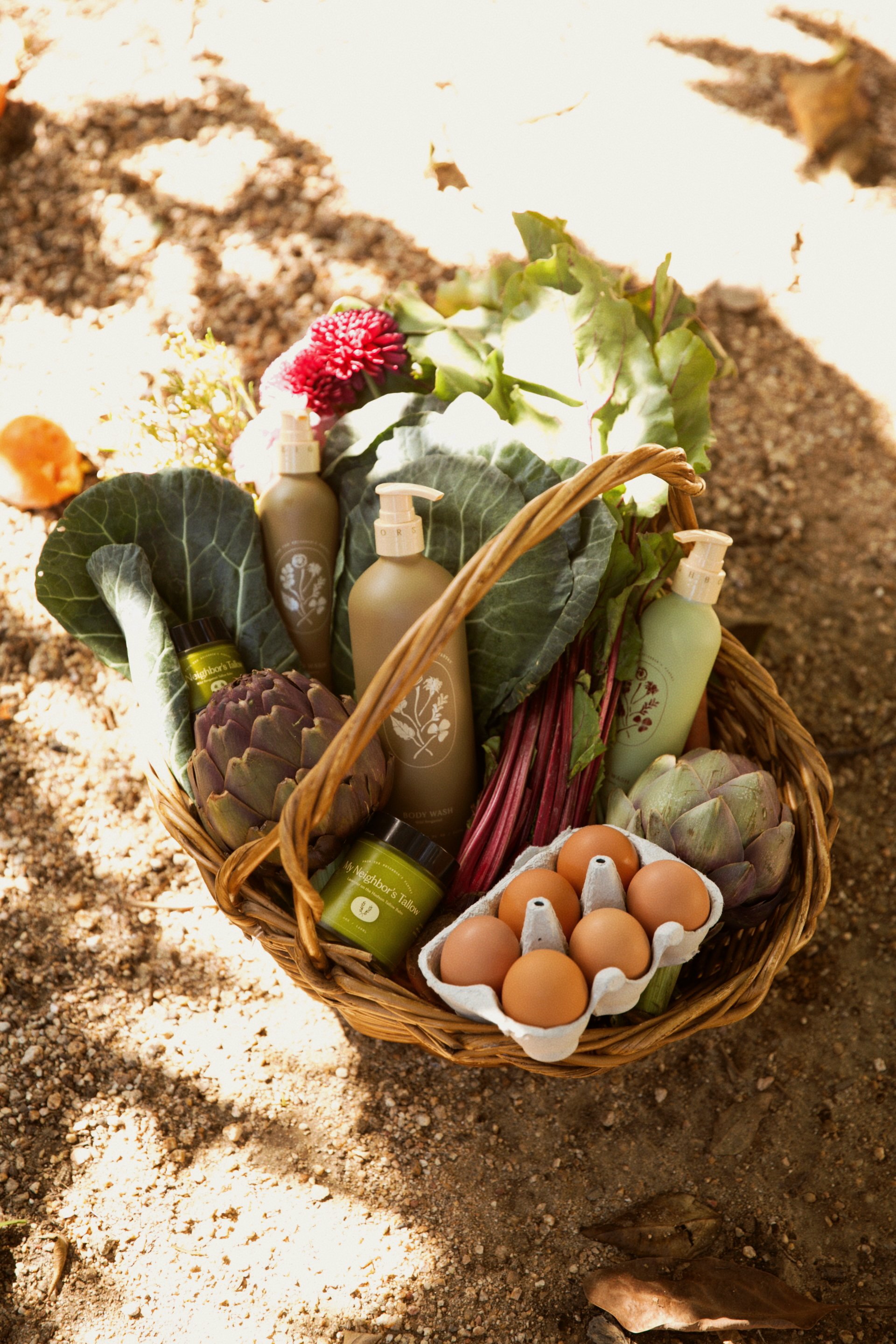 A wicker basket filled with eggs, artichokes, leafy greens, herbal products, and a pink flower, placed on sandy ground with leaves scattered around.