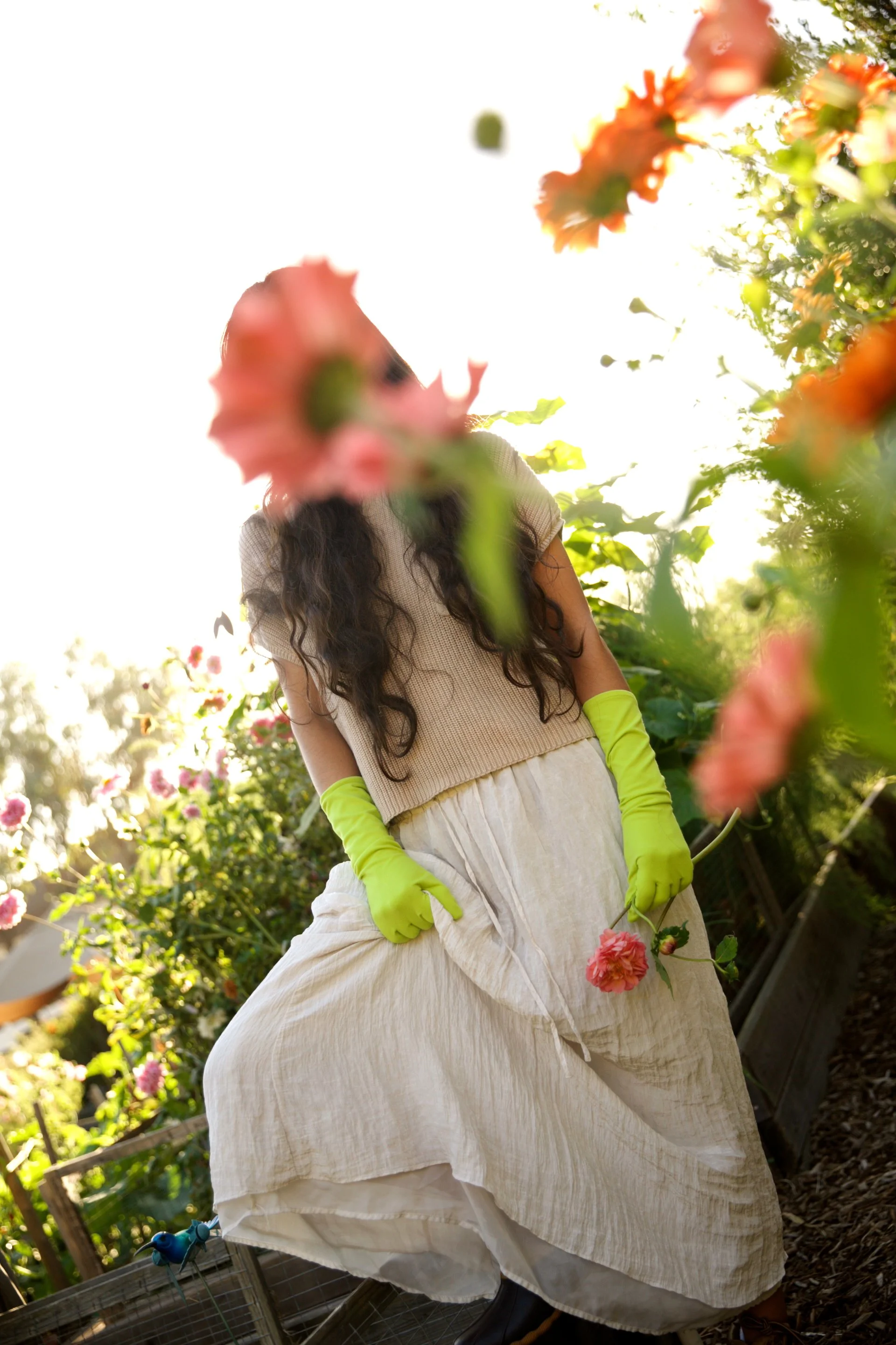A woman with long dark curly hair wearing a beige top, a long white skirt, bright yellow-green gloves, and black boots standing in a garden. She is partially obscured by pink flowers in the foreground, holding a pink flower in her hand, with sunlight