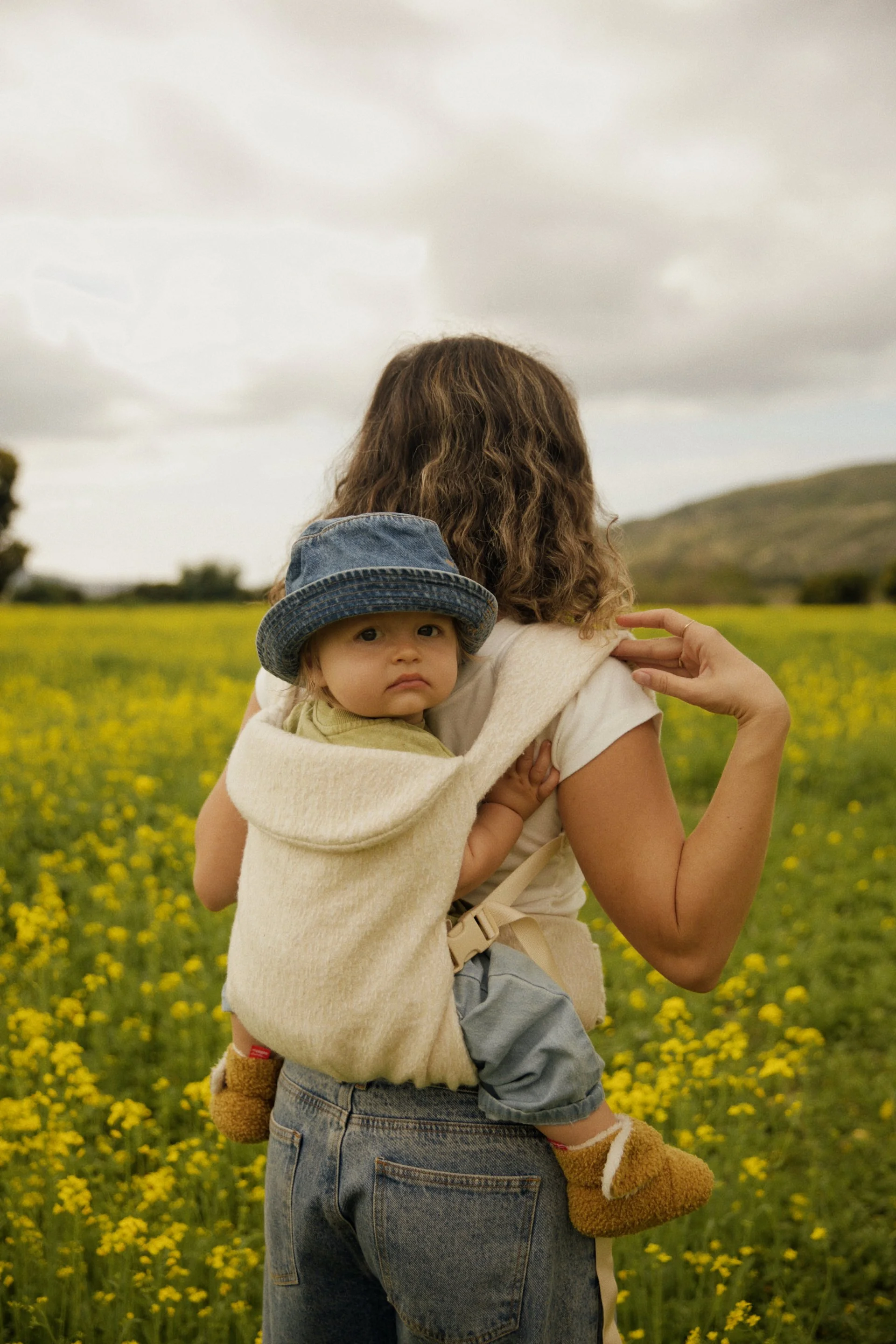 A woman holding a young child in a yellow flower field with a hill in the background, cloudy sky overhead.