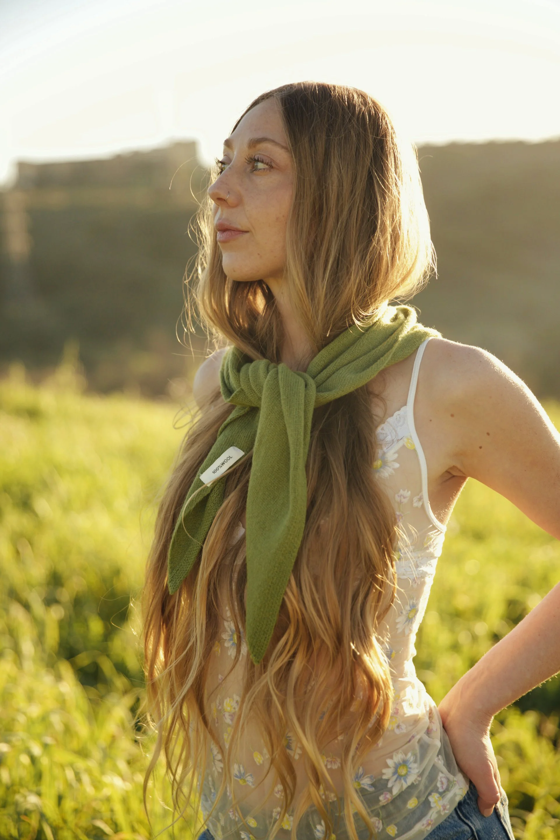 A young woman with long, wavy red hair in a field during golden hour, wearing a white tank top with a floral pattern, denim shorts, and a green scarf tied around her neck, gazing to her left with a soft expression.