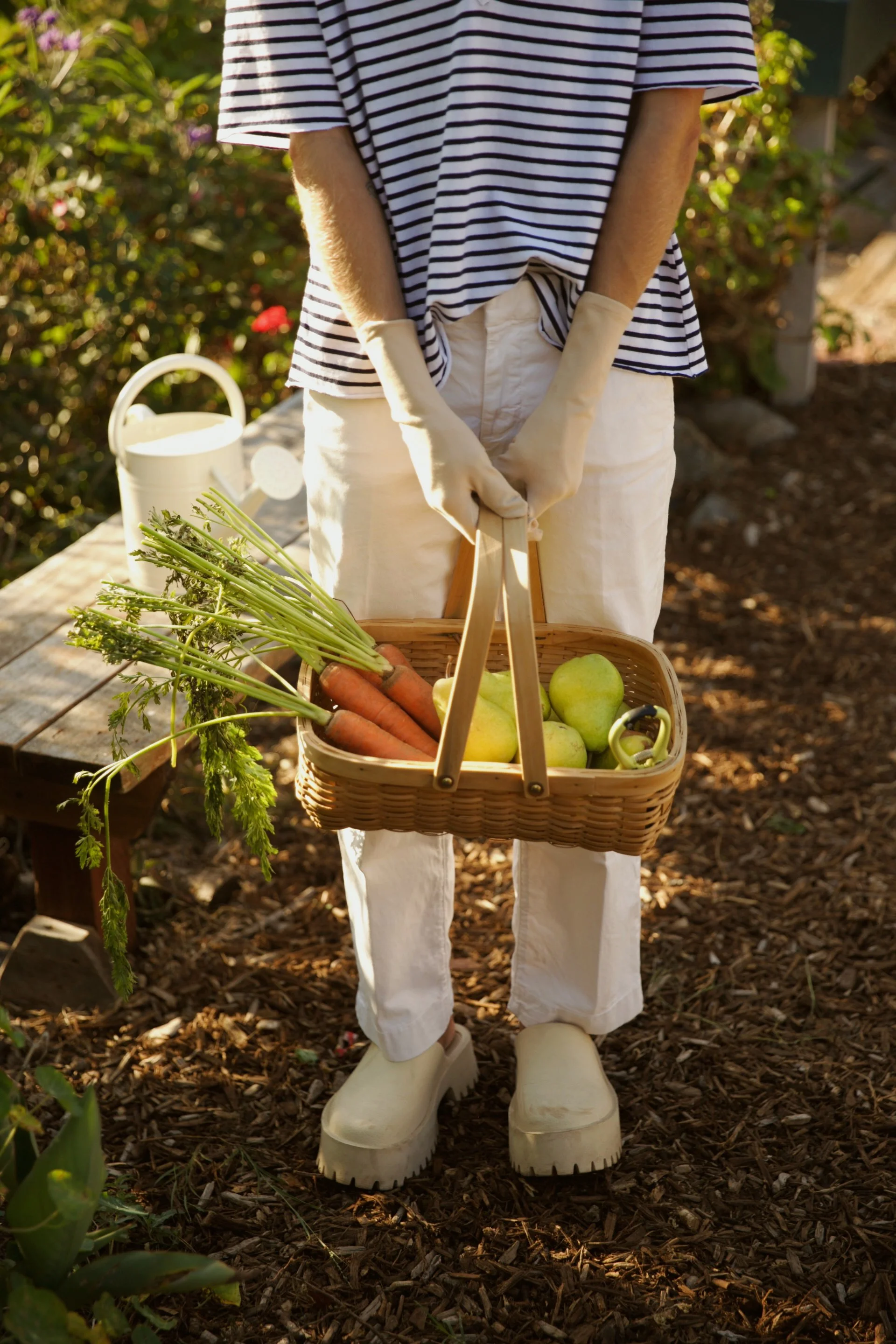 A person wearing white gloves and cream-colored boots holding a basket of fresh vegetables, including carrots and apples, in a garden.