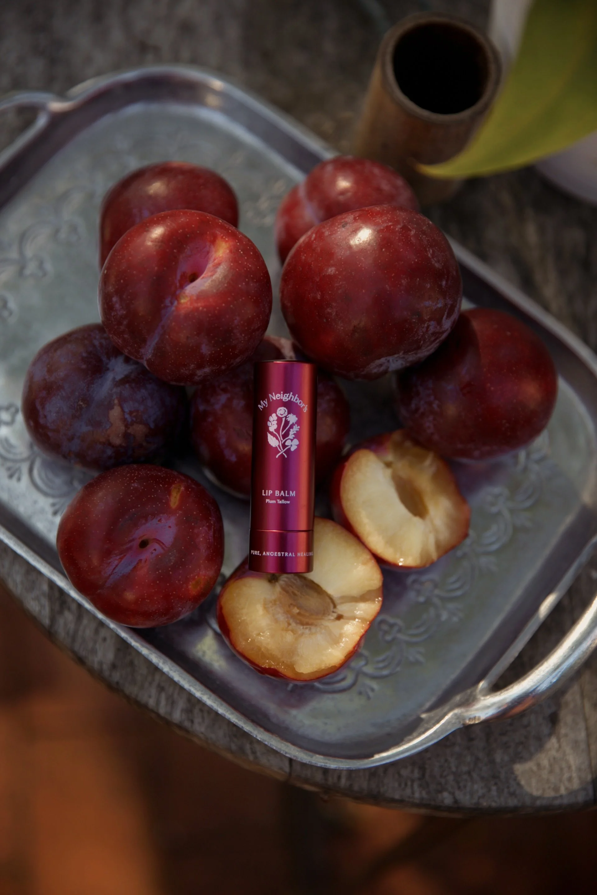 A silver tray with plums, some sliced showing their white interior and brown seeds, and a red lip balm stick labeled 'My Neighbors' Lip Balm Plum Tallow' placed among the apples.