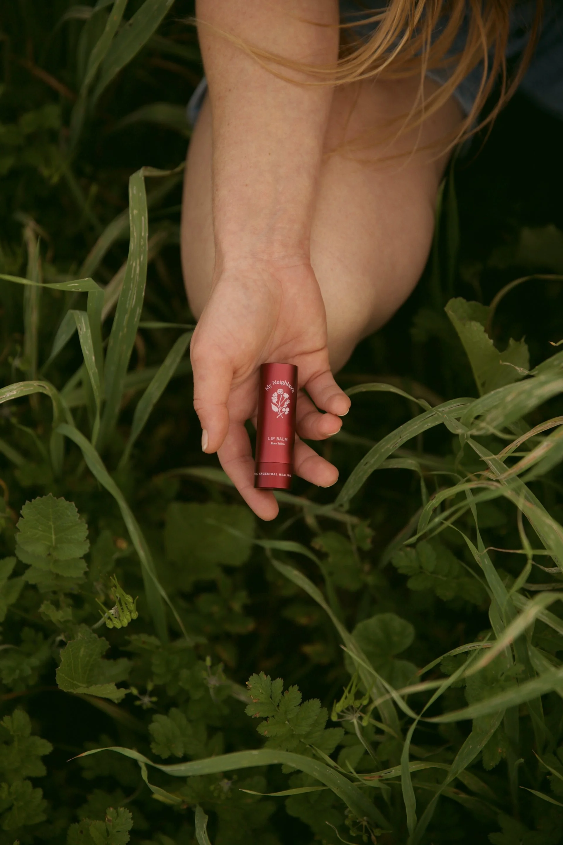 A person holding a red lipstick tube labeled 'My Neighbors Lip Balm' in a grassy outdoor setting.