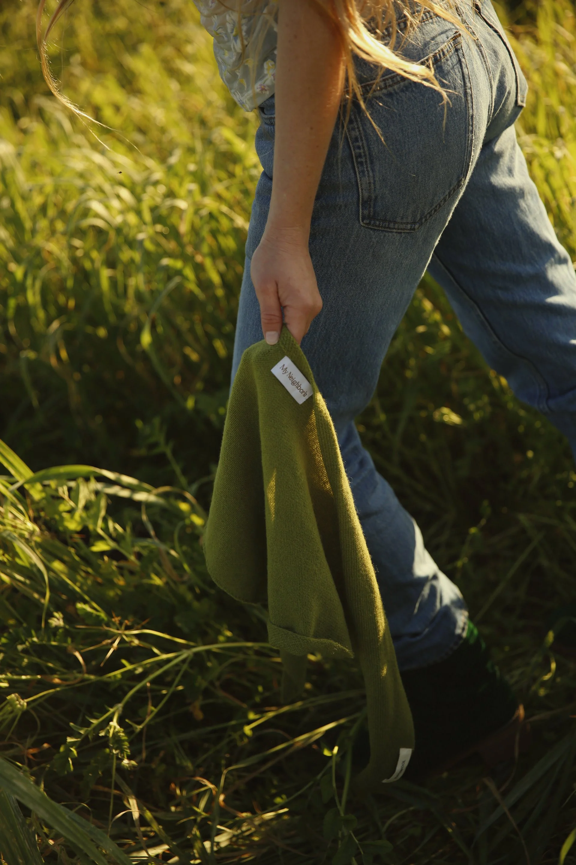 A person wearing jeans and a floral shirt is bending down in a field of tall grass, holding a green piece of cloth with a white label that reads "My Neighbors."