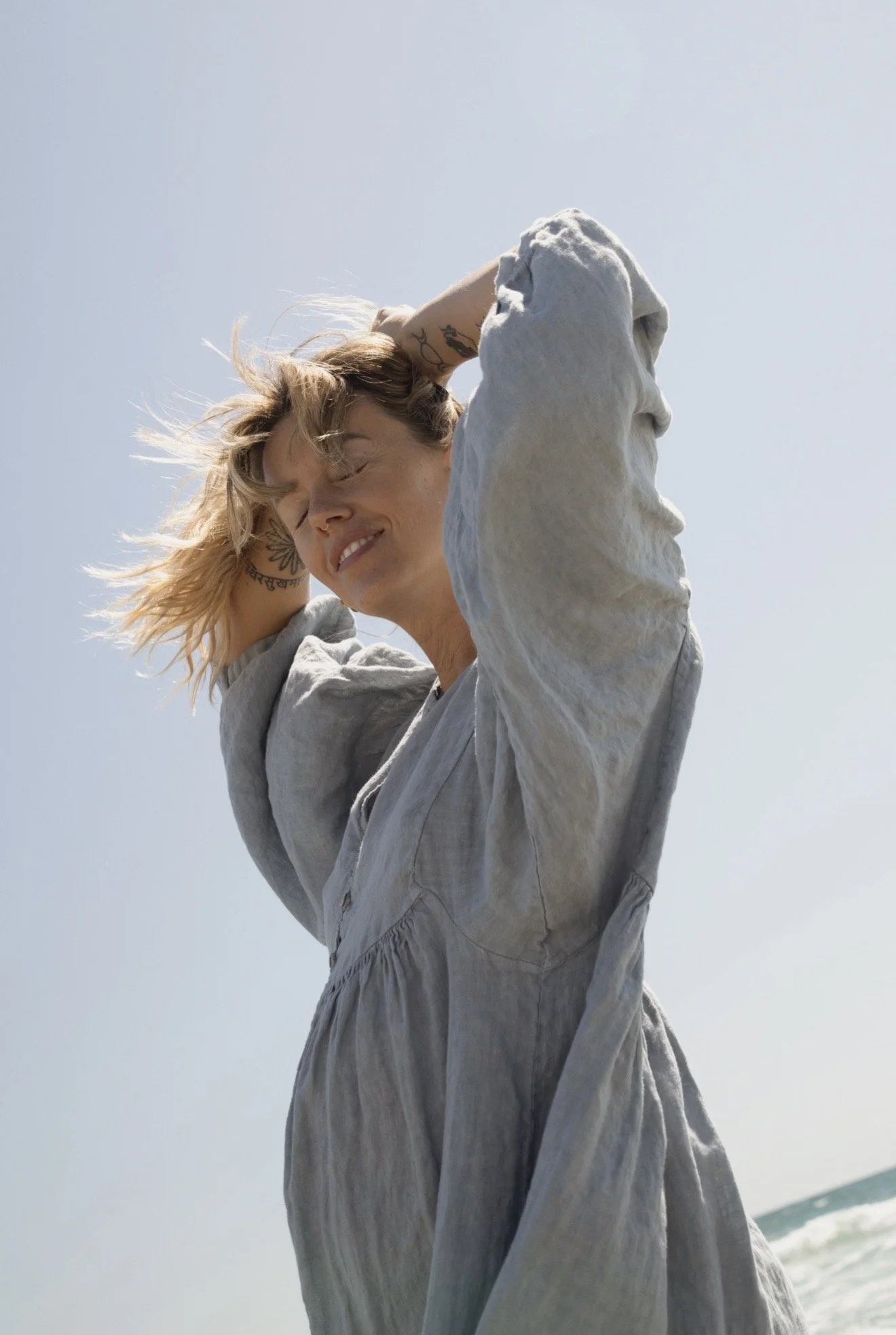 A woman with blonde hair, tattoos, and a nose ring standing outdoors in front of a bright sky, smiling with eyes closed, wearing a light-colored dress or cover-up.