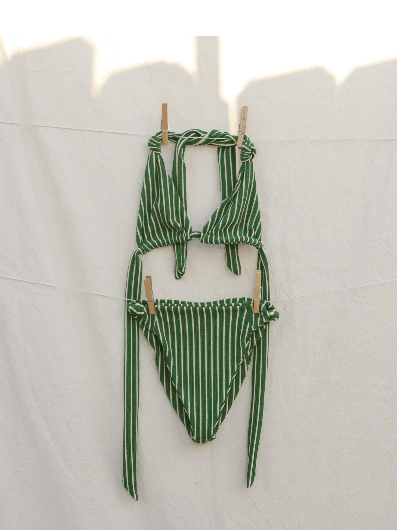 Green and white striped bikini top and bottom hanging on a clothesline with clothespins against a white wall.