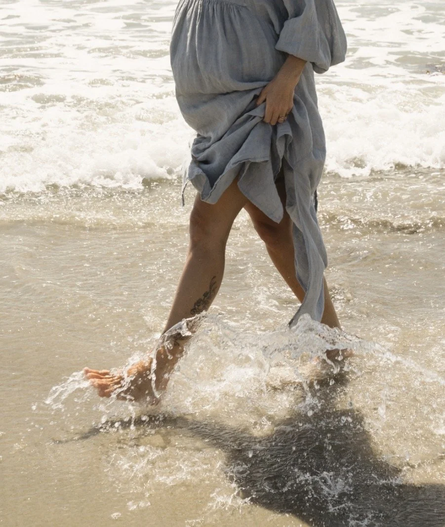 Person wearing a gray dress walking barefoot in shallow ocean water.