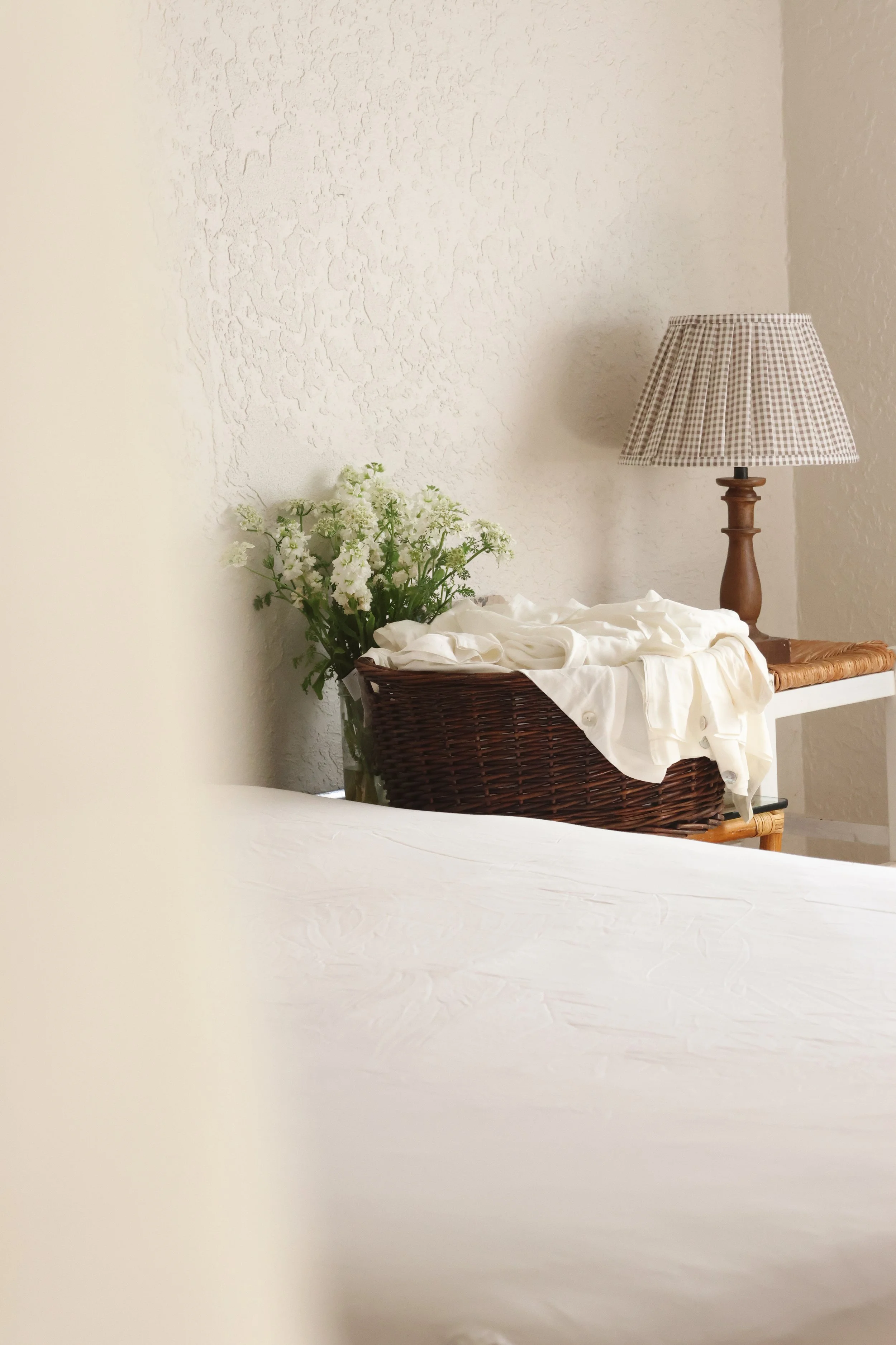 A wicker basket filled with white fabric, a bouquet of white flowers, a wooden table, and a table lamp with a checked fabric shade against a textured off-white wall.