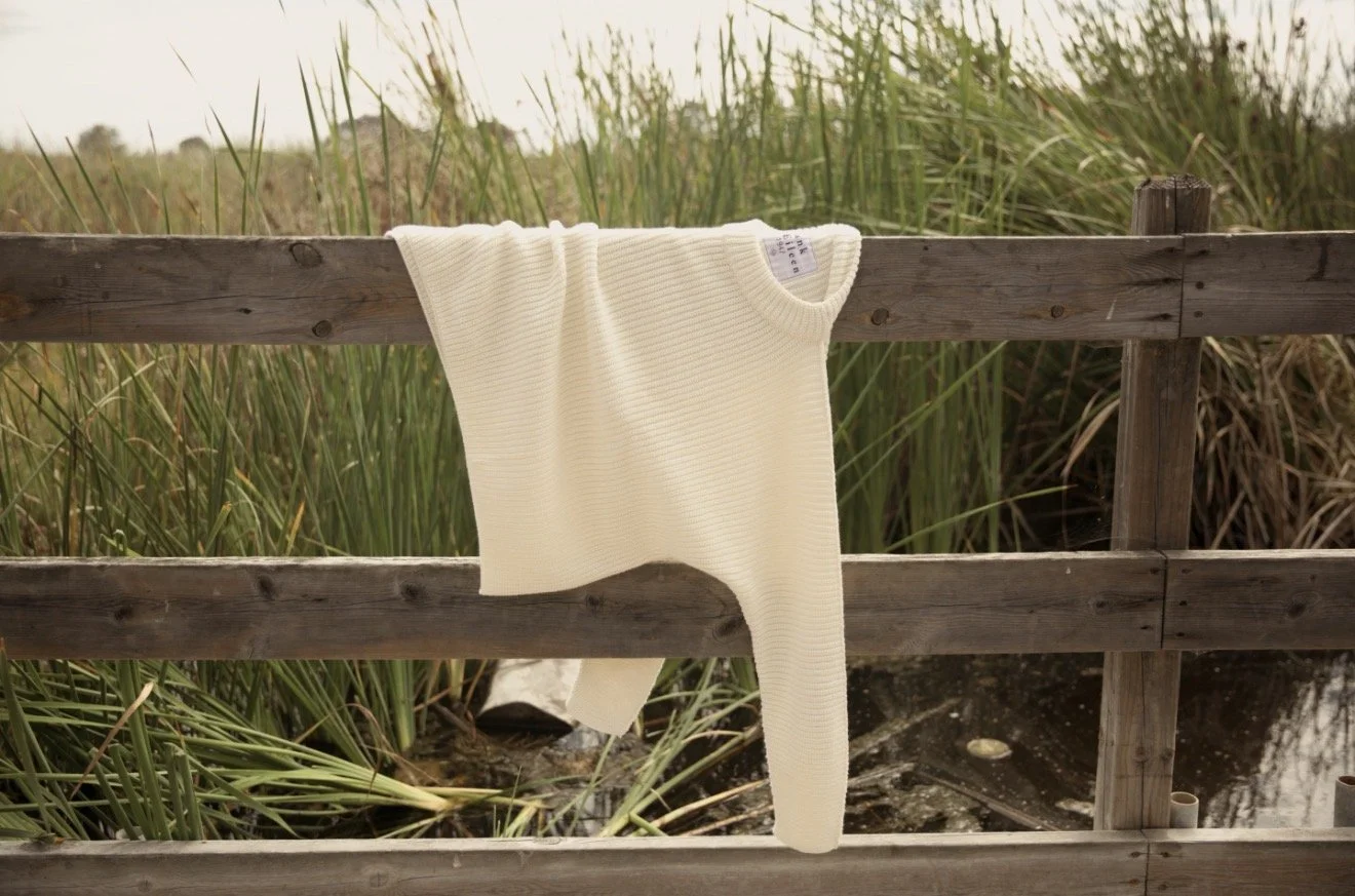 A cream-colored sweater hanging over a wooden railing with green reeds and a pond in the background.