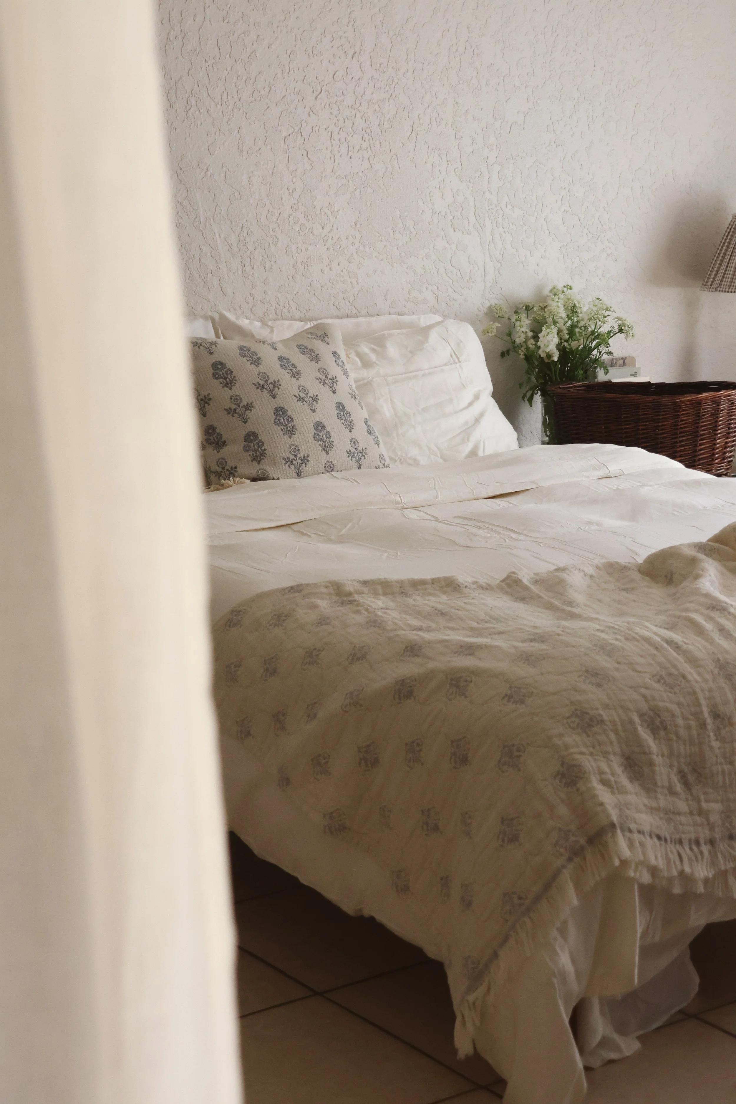Part of a bed with white and patterned pillows, a cream-colored bedspread, a wicker basket with white flowers, and a wall with textured paint.