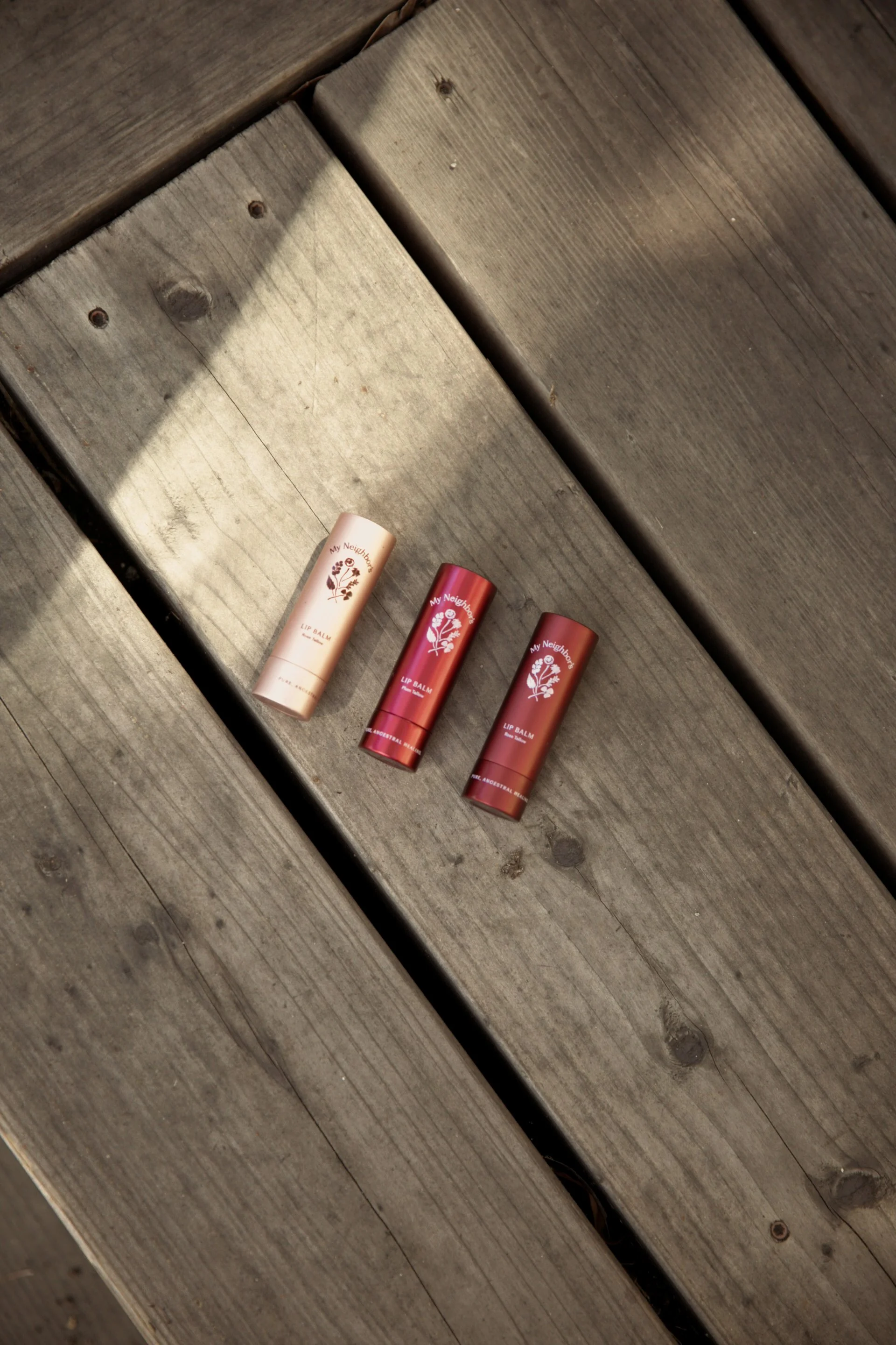 Three tubes of lip balm resting on a weathered wooden surface. The lip balms are in beige, red, and brown containers, labeled with a floral design and text, arranged horizontally.