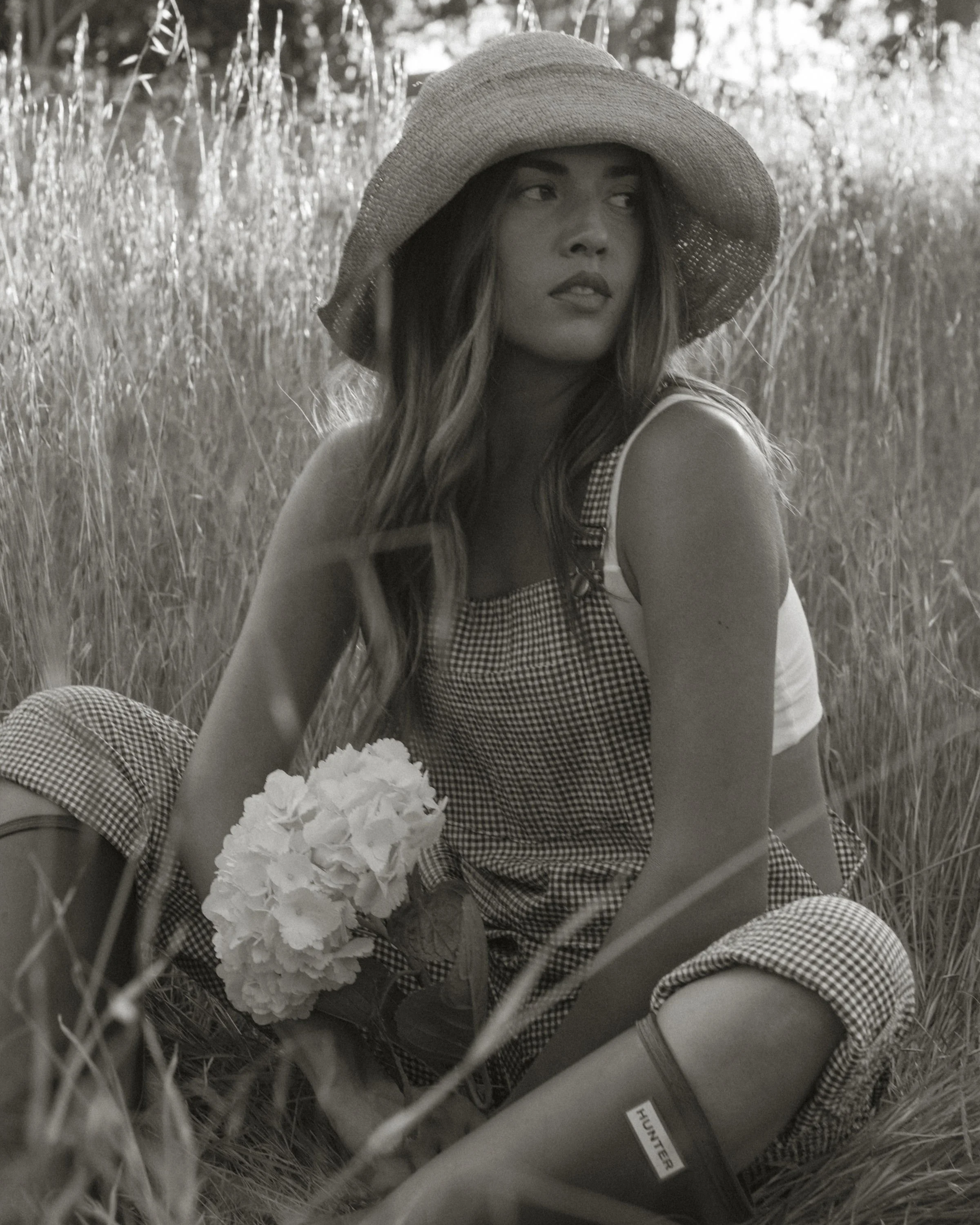 A woman sitting in a field of tall grass, wearing a large sun hat and a checkered dress, holding a bouquet of flowers.