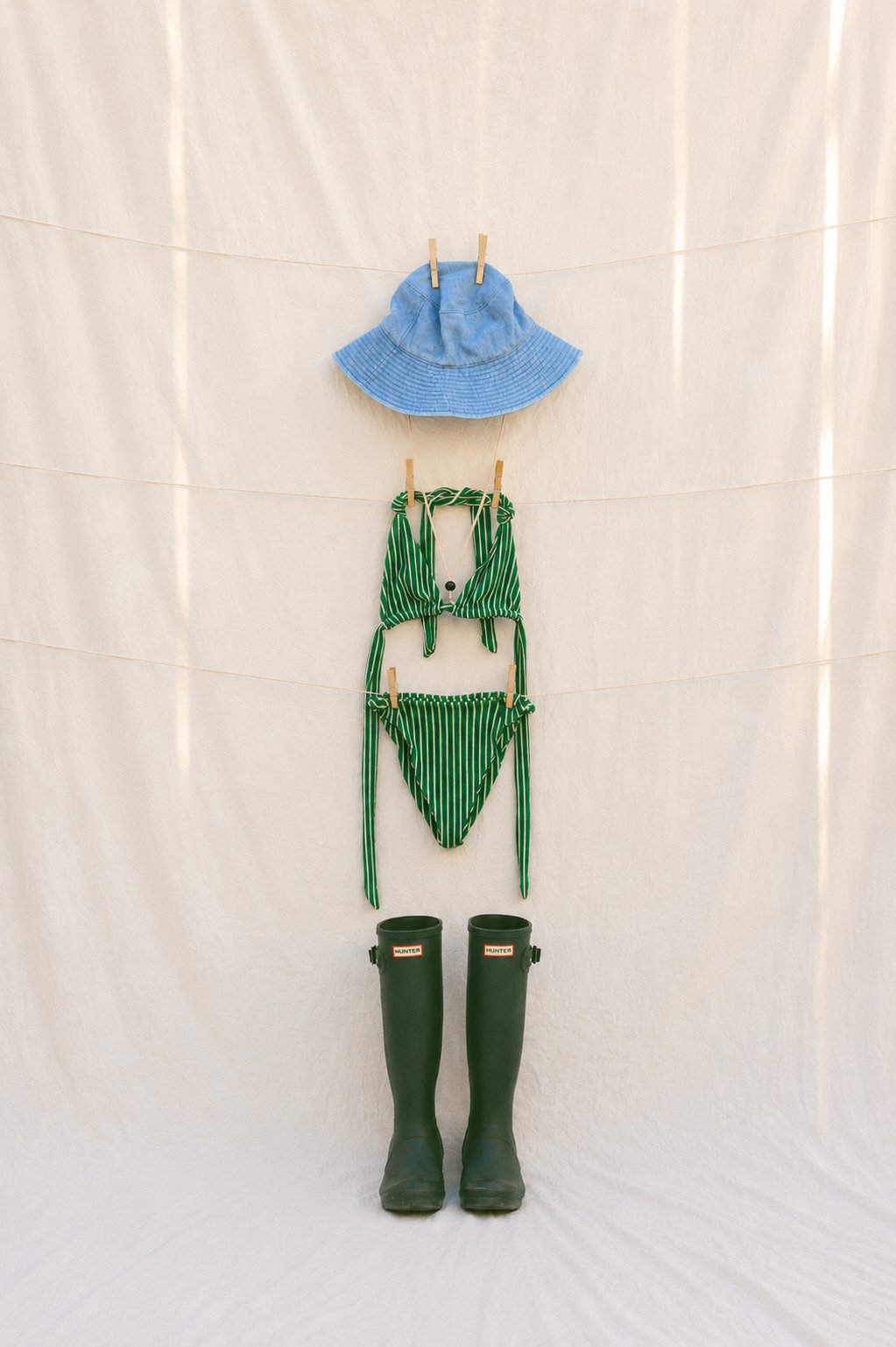 A blue sun hat, a green and white striped bikini, and a pair of green rubber rain boots hanging on a beige fabric backdrop.