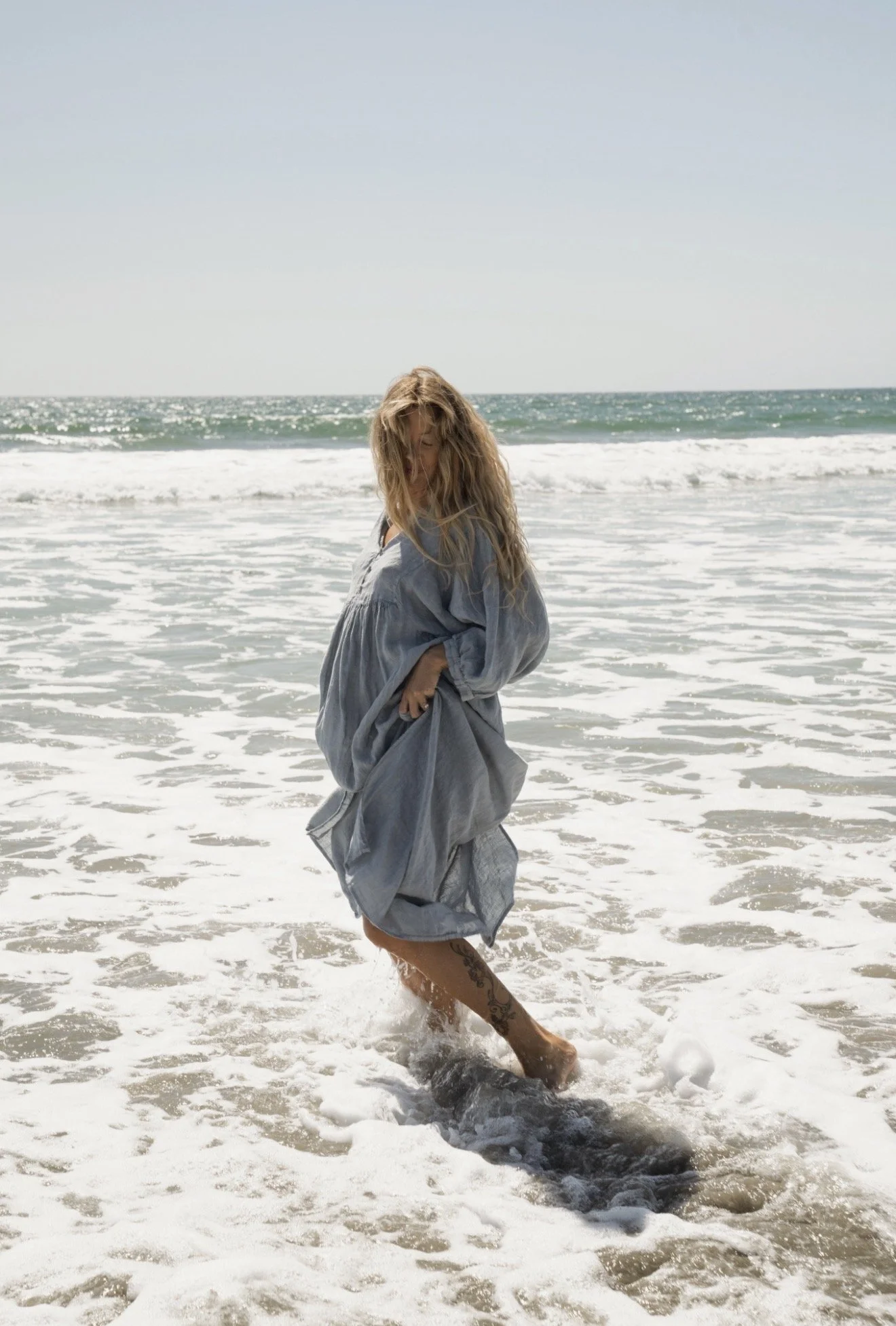 A woman with long blonde hair is walking in the ocean surf, wearing a light blue dress, with waves and a clear sky in the background.