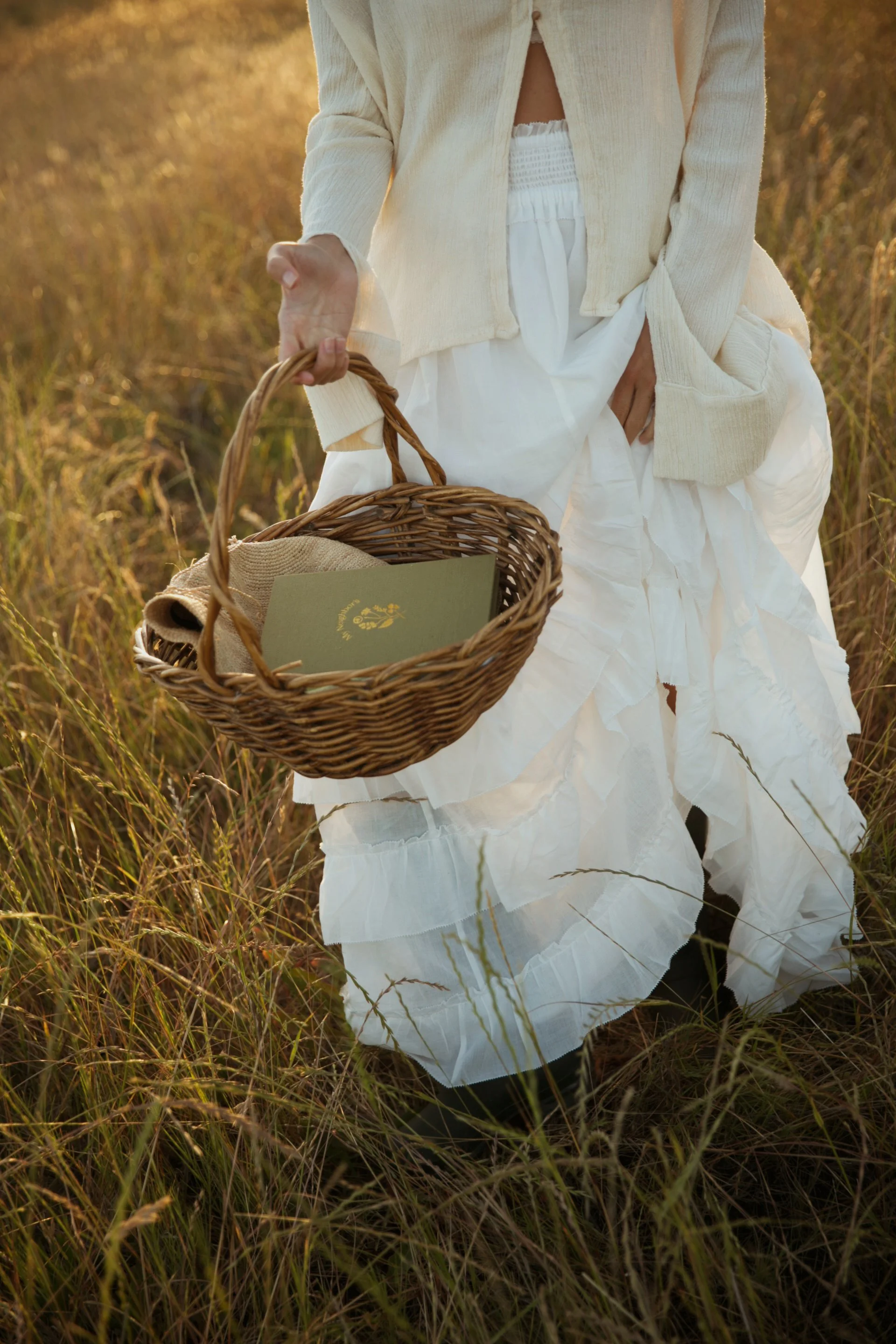 Person in cream-colored cardigan and white dress holding a wicker basket in a field of tall grass during sunset.
