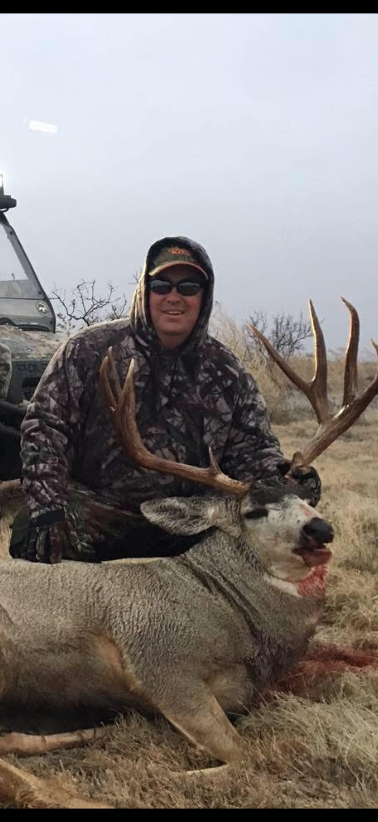 A person in camouflage clothing and sunglasses kneeling behind a large deer with antlers, which is lying on the ground in a grassy outdoor area.