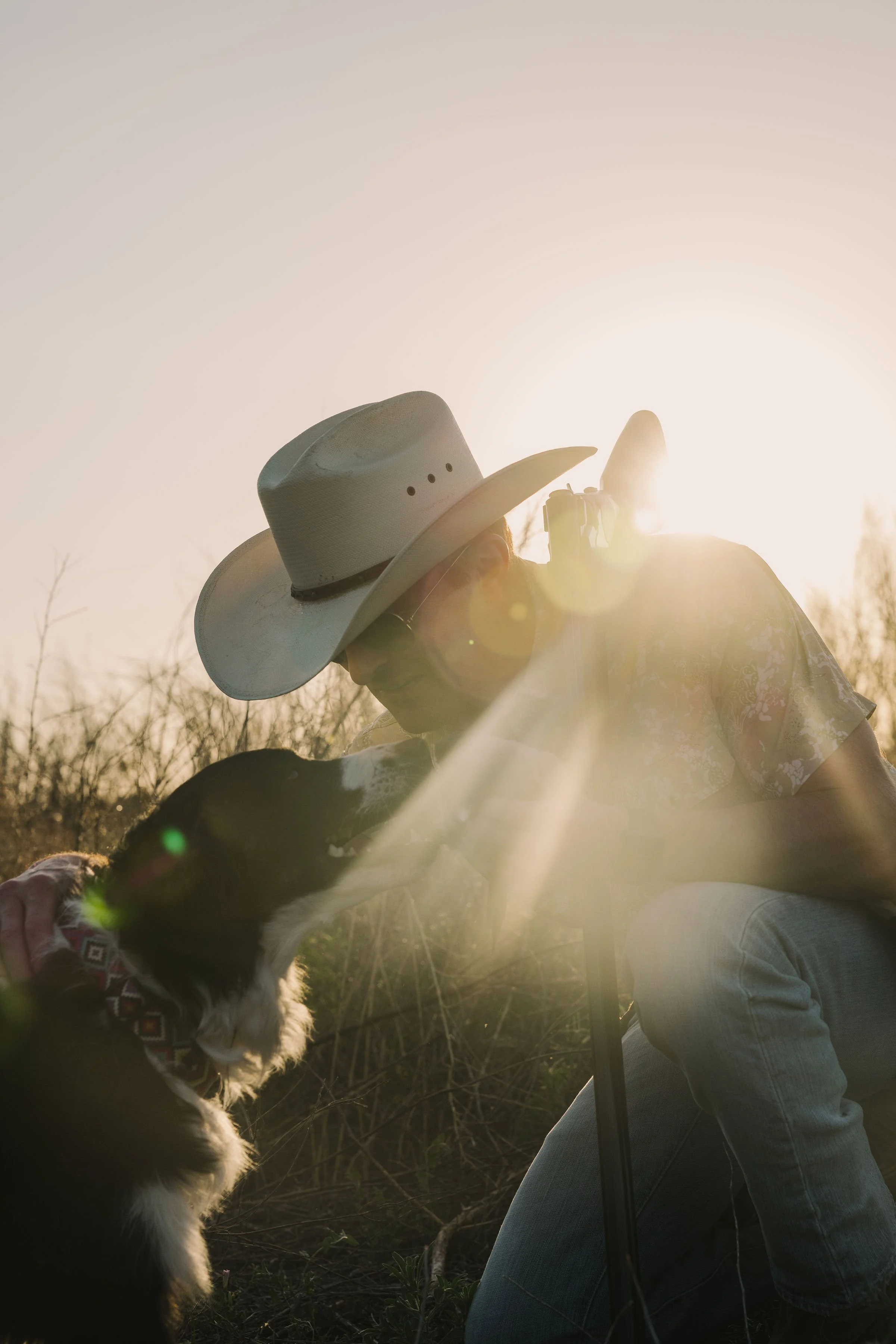 Person wearing a cowboy hat and sunglasses sitting outdoors with a dog during sunset.