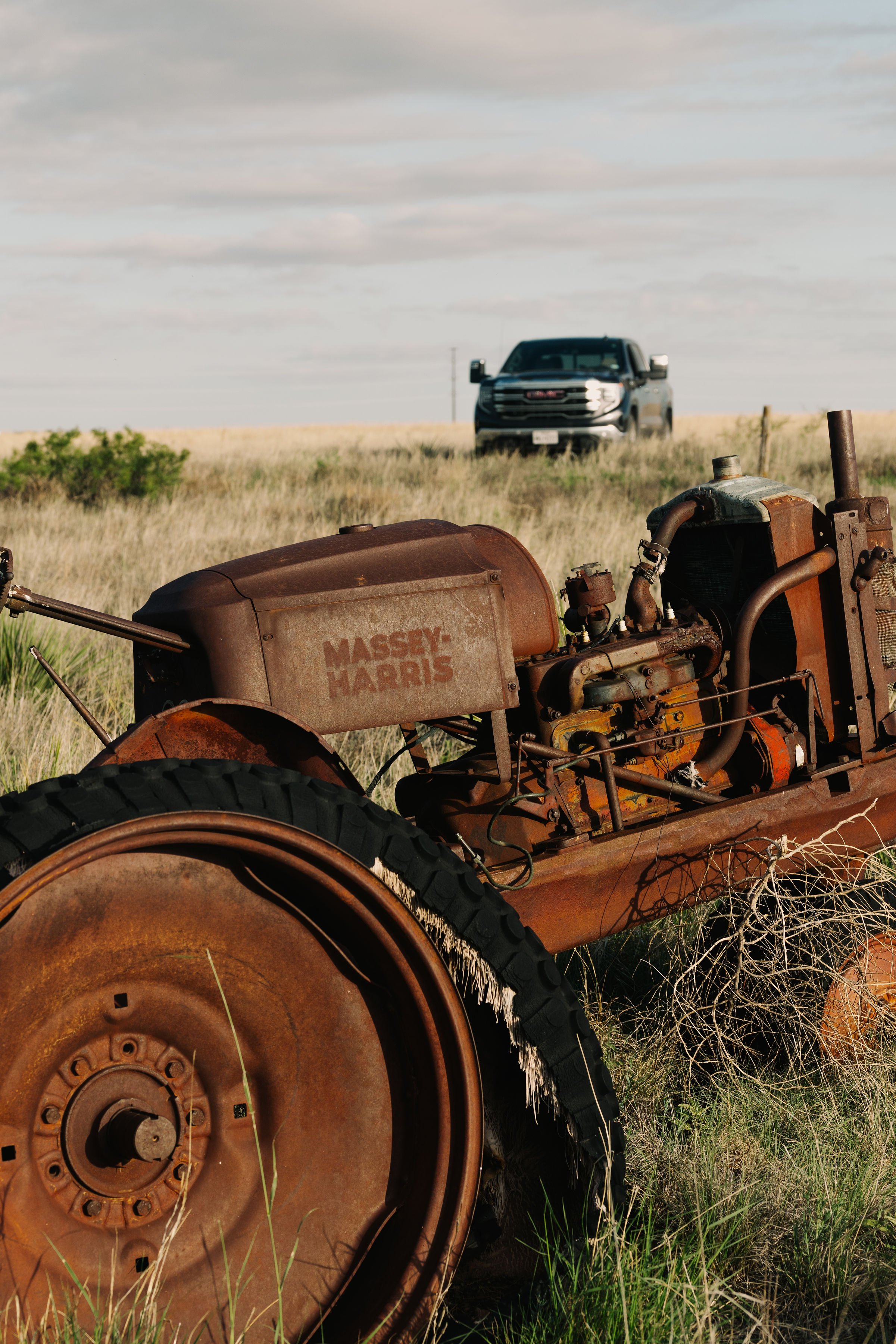 Rusty tractor in grassy field with an old GMC pickup truck in the distance under a cloudy sky