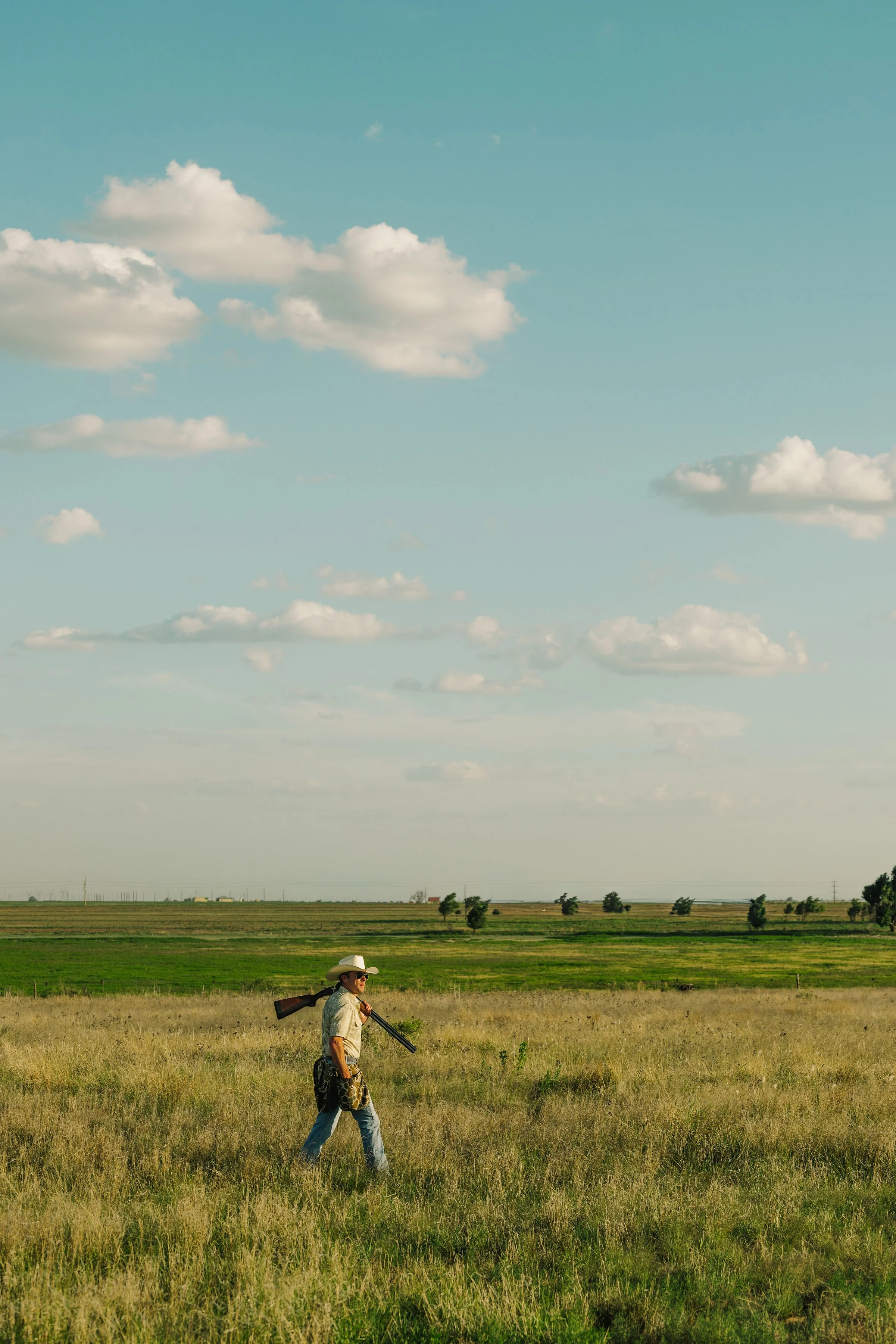 A person wearing a cowboy hat, sunglasses, and patterned pants walking through a grassy field while carrying a rifle over their shoulder. The landscape features green fields with scattered trees and a partly cloudy sky.