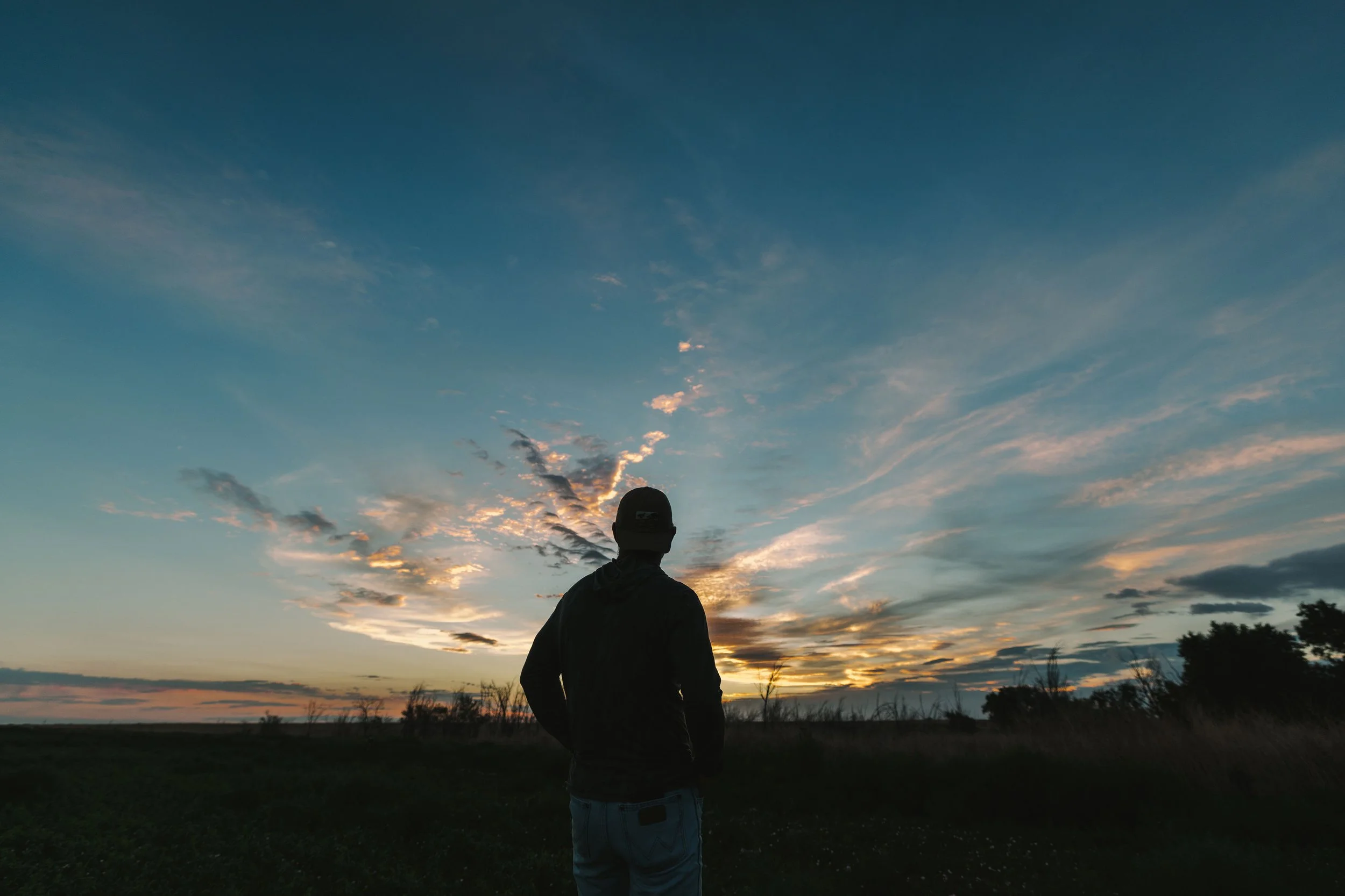A person standing outdoors at dusk, facing away from the camera, with a vast sky filled with clouds and a setting sun on the horizon.