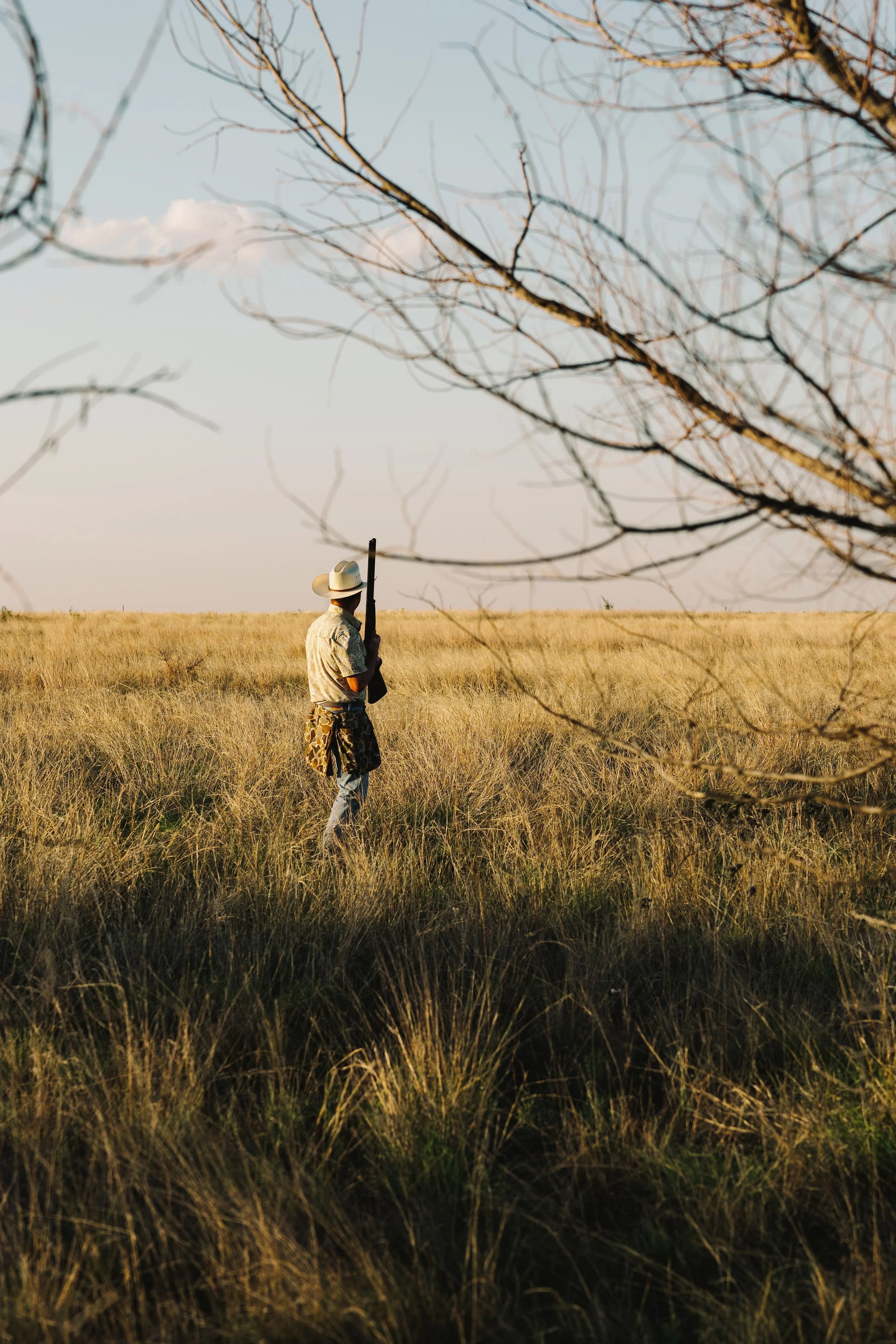 Man in camouflage pants and a hat holding a rifle in a field with tall grass and leafless trees, sunset or sunrise lighting creating a warm glow.