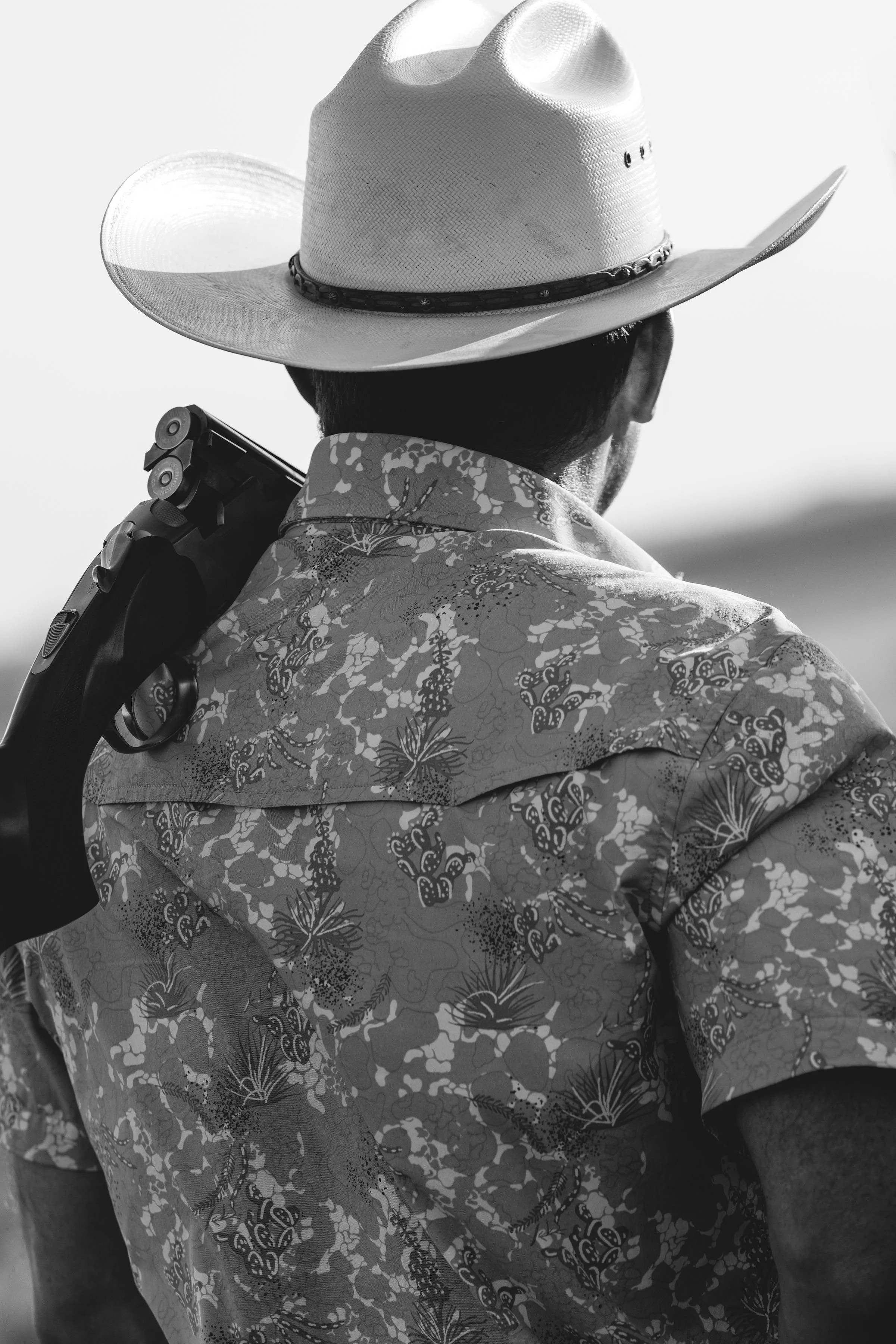 A person wearing a cowboy hat, patterned shirt, and carrying a gun on their shoulder, viewed from behind, in black and white.