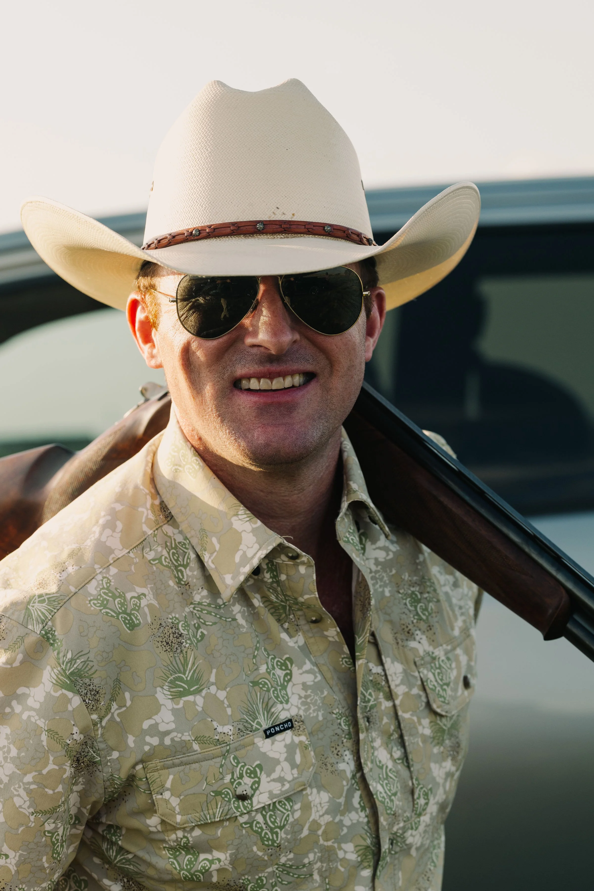 A man wearing a white cowboy hat, dark sunglasses, a patterned beige shirt with green accents, and carrying a rifle over his shoulder.