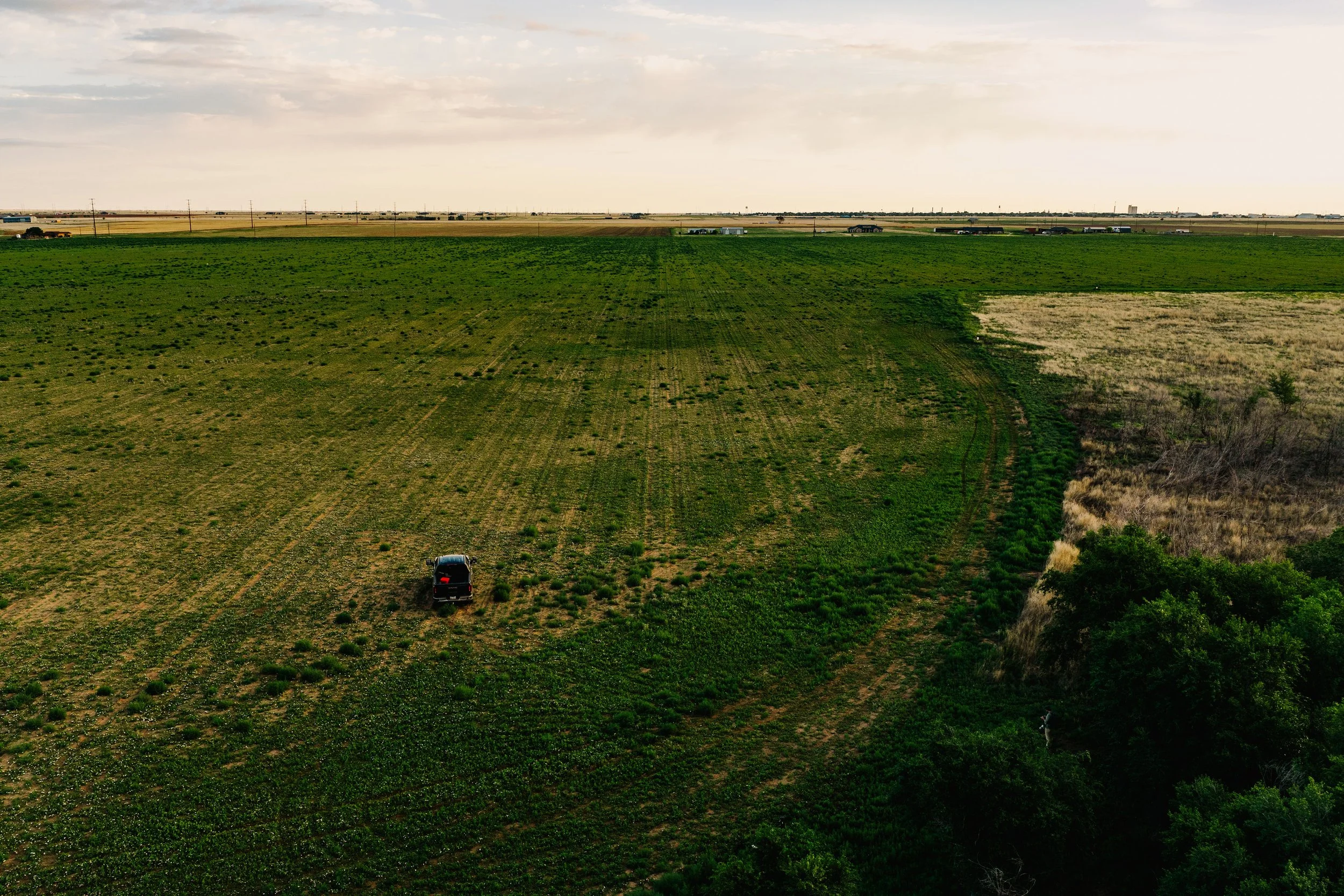 A wide view of a rural area with green farmland and sparse trees. There is a single black vehicle parked on the farmland. The background shows a distant road with multiple cars and trucks driving along it. The sky is partly cloudy with sunlight illuminating the scene.