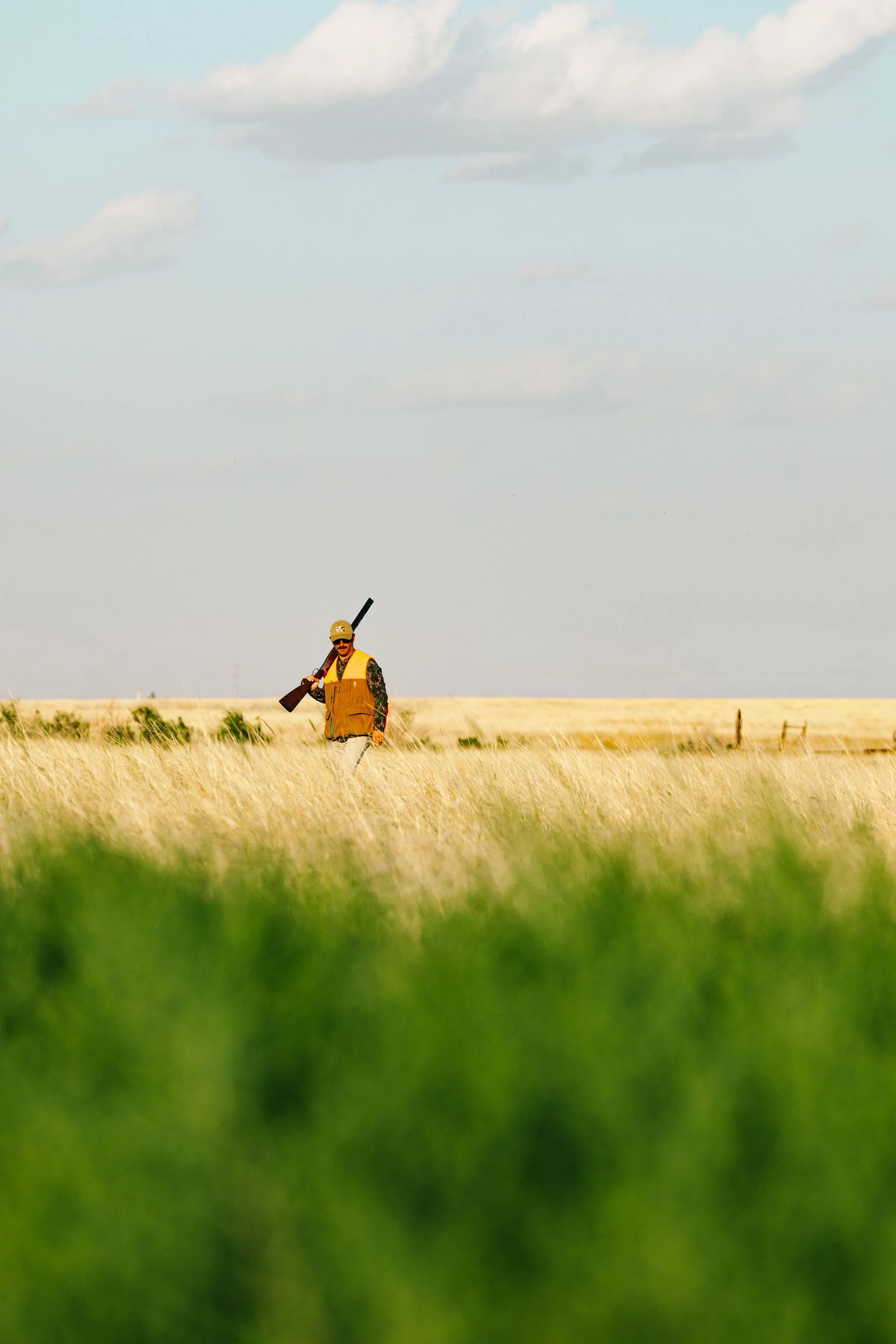 A man walking through a grassy field while carrying a shotgun over his shoulder under a cloudy sky.