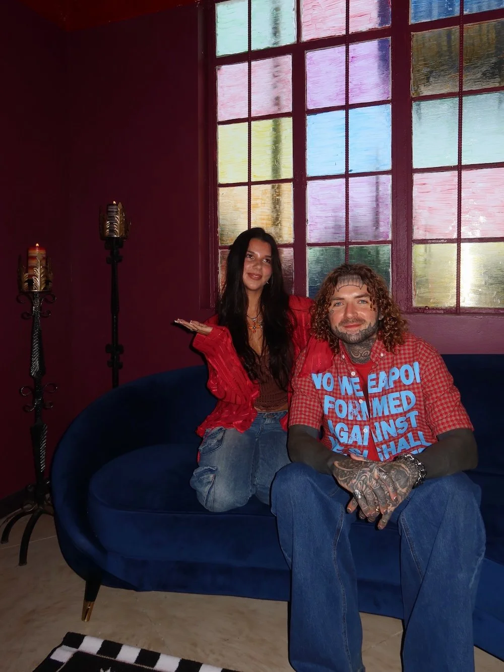 A woman and a man sitting on a dark blue couch in front of a colorful window with stained glass panels. The woman is wearing a red jacket and distressed jeans, sitting cross-legged. The man has curly hair, tattoos, and is wearing a red plaid shirt with blue text, sitting with his hands clasped. The room has dark red walls and candle holders.